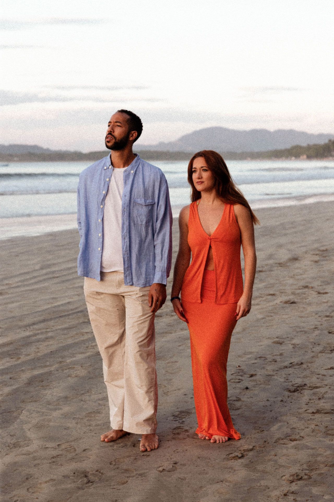 A couple walking together on a sandy beach at sunset wearing a blue shirt and orange dress.