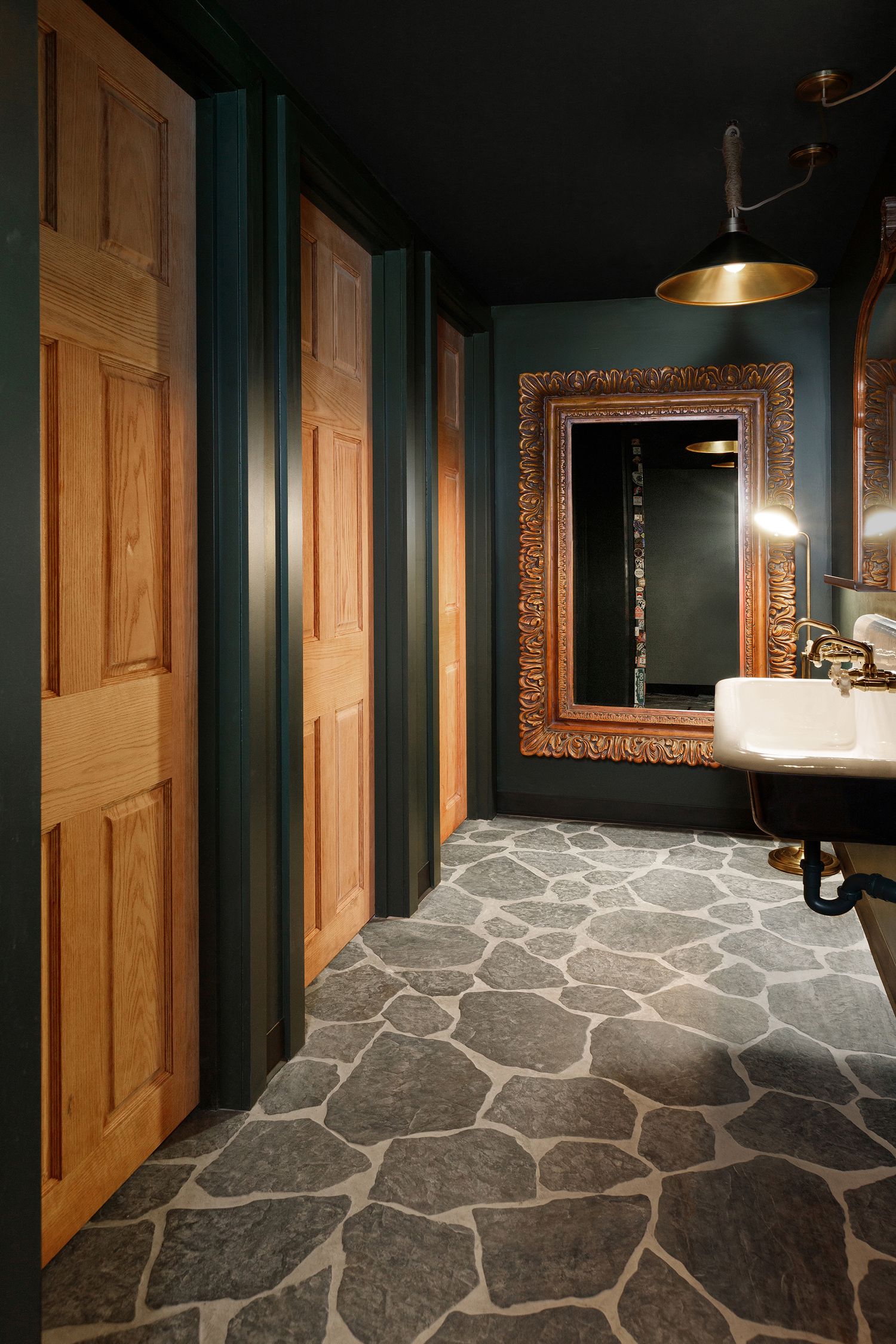 Rustic bathroom with stone floor, dark teal walls, wood doors, ornate gold mirror, and vintage white sink.