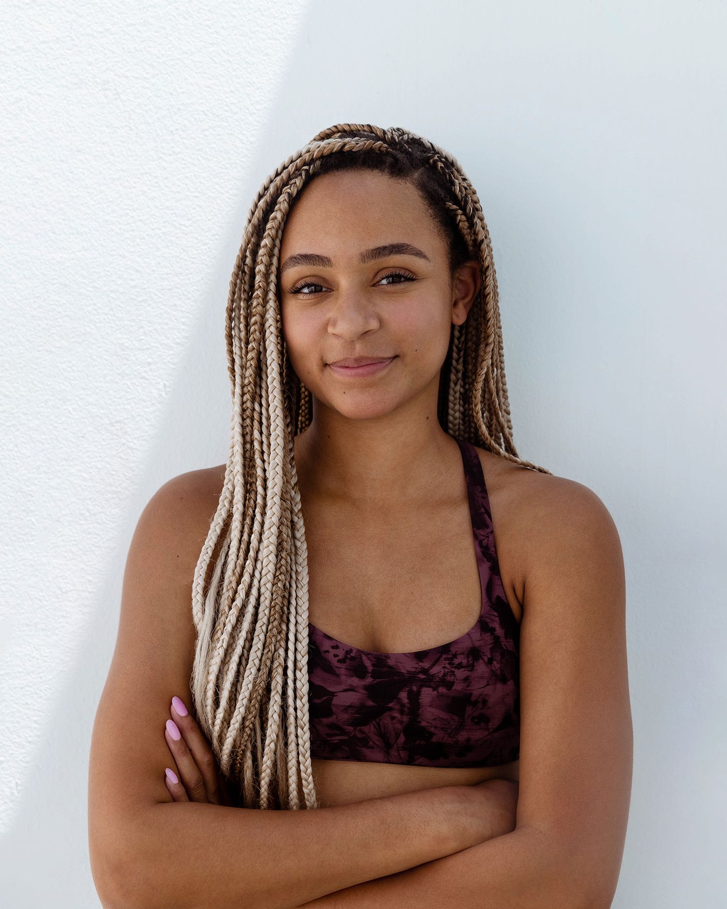 Woman with blonde braids wearing a purple floral top poses against white background.