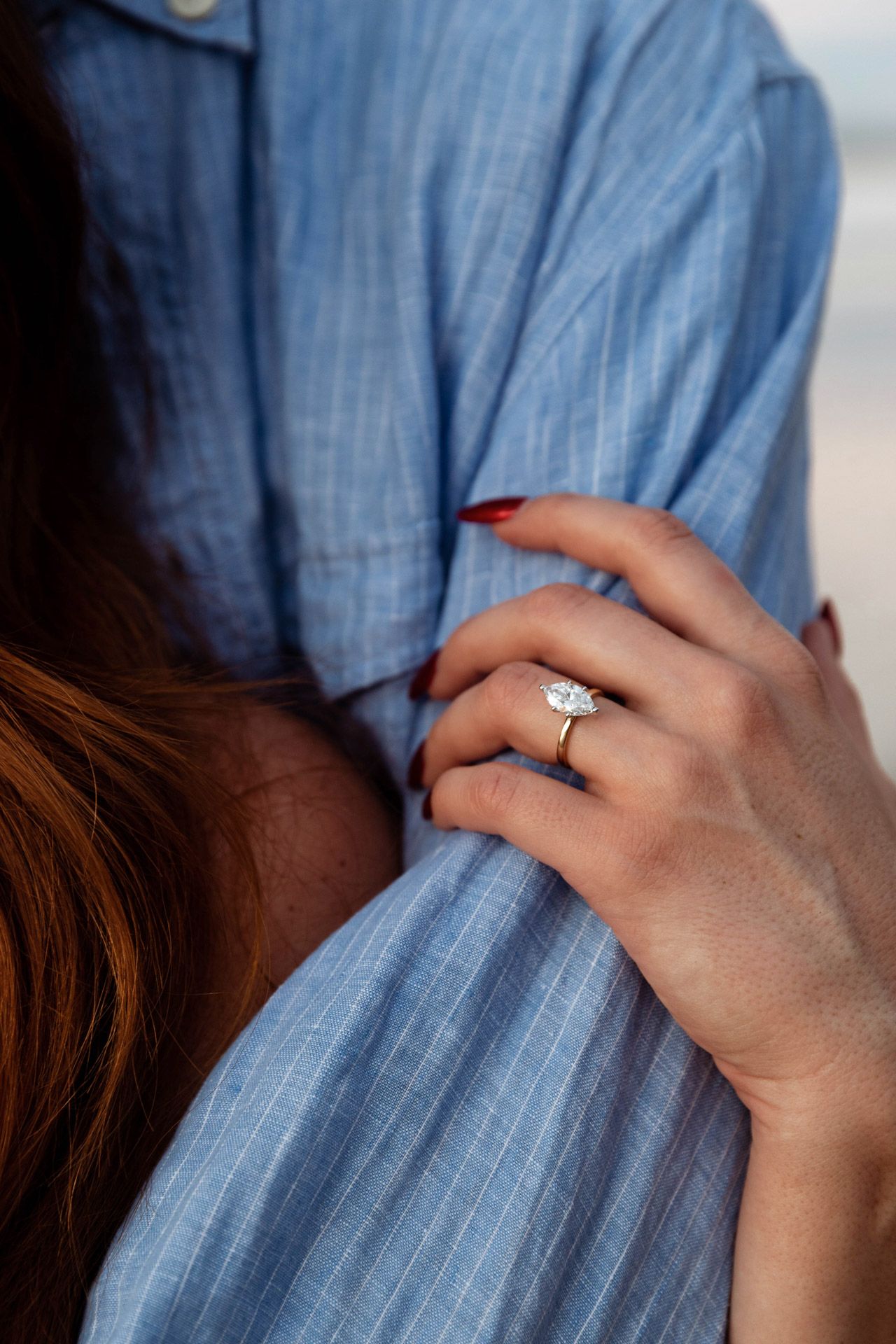 Close up of an engagement ring on a hand gripping a blue striped shirt against an ocean backdrop.