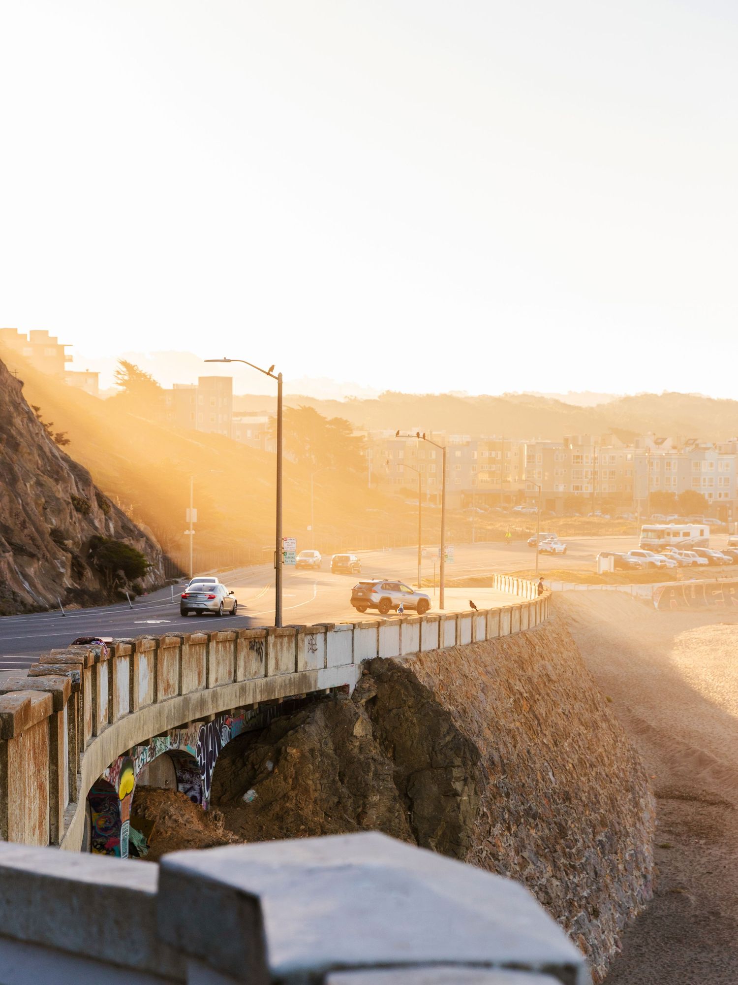 Golden sunset light illuminating a curved coastal highway bridge with cars and city buildings beyond.