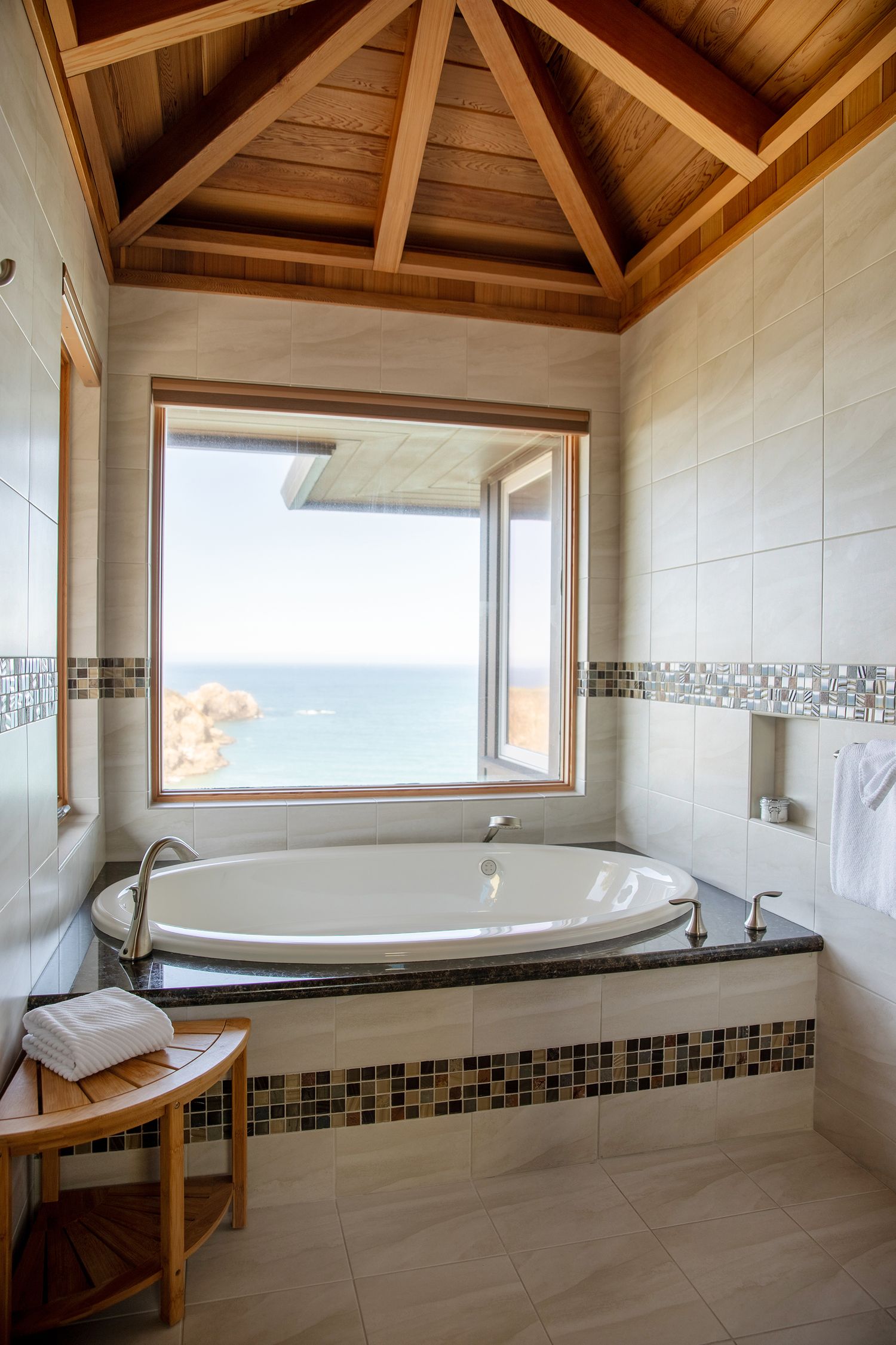 Luxurious bathroom with oval soaking tub under vaulted wood ceiling featuring large window with ocean view.