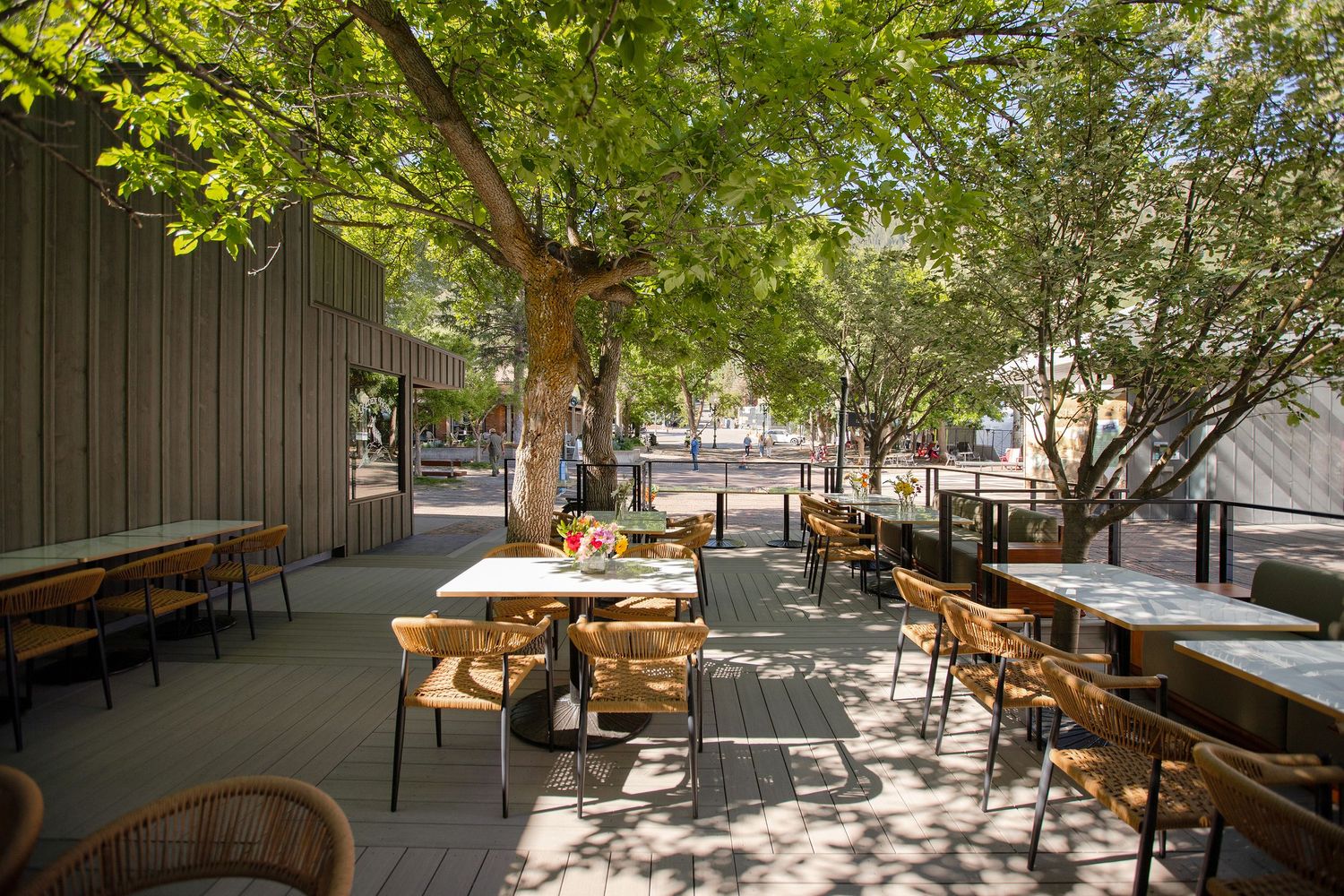 Shaded outdoor patio dining area with wicker chairs, white tables, and mature trees providing natural canopy.