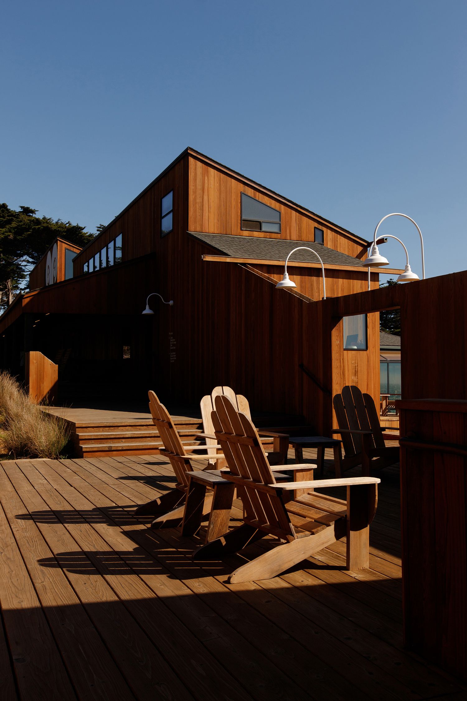 Adirondack chairs on a wooden deck outside a modern cedar-clad building under a clear blue sky.