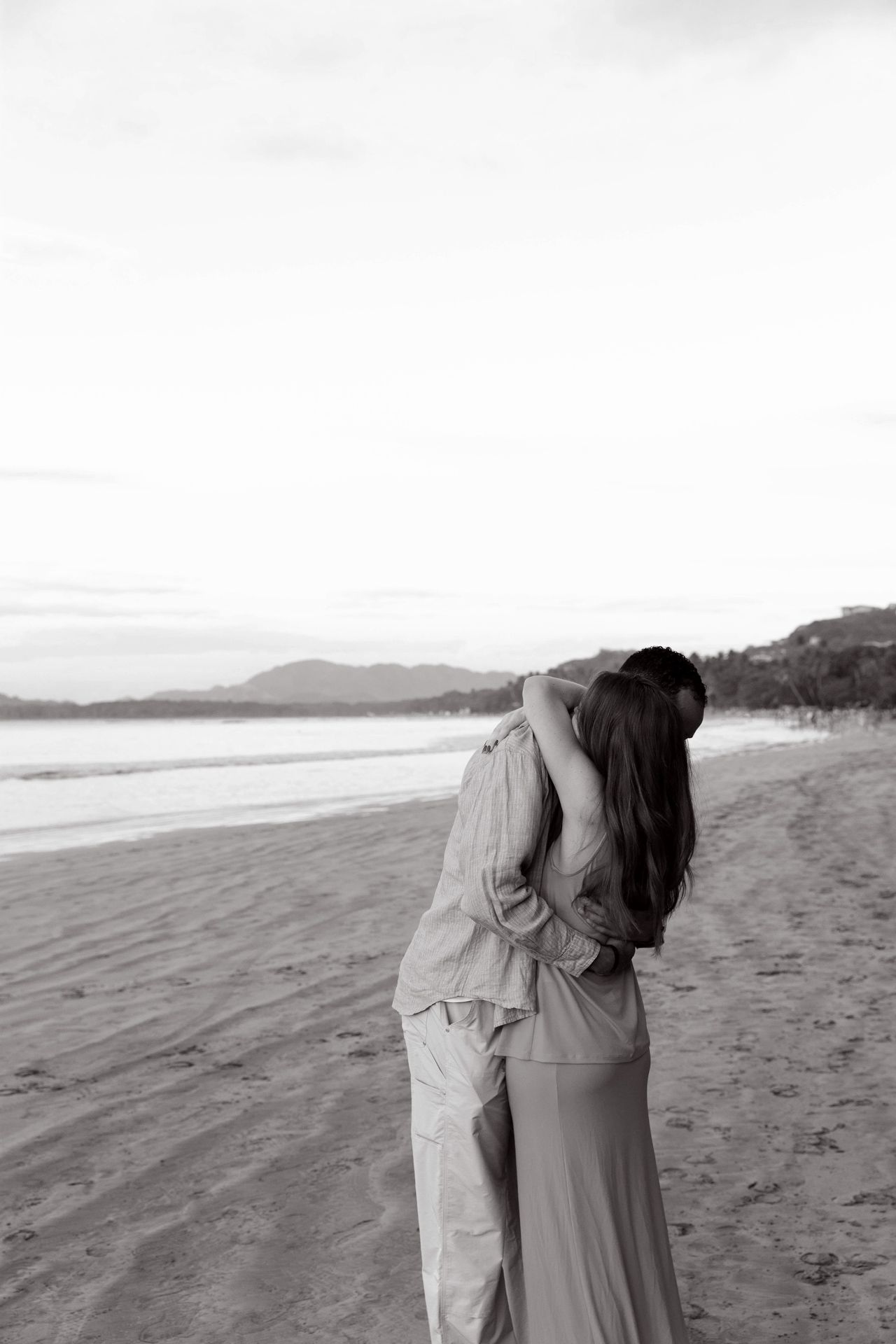 A couple embraces on a scenic beach with mountains in the distance during a black and white photoshoot.