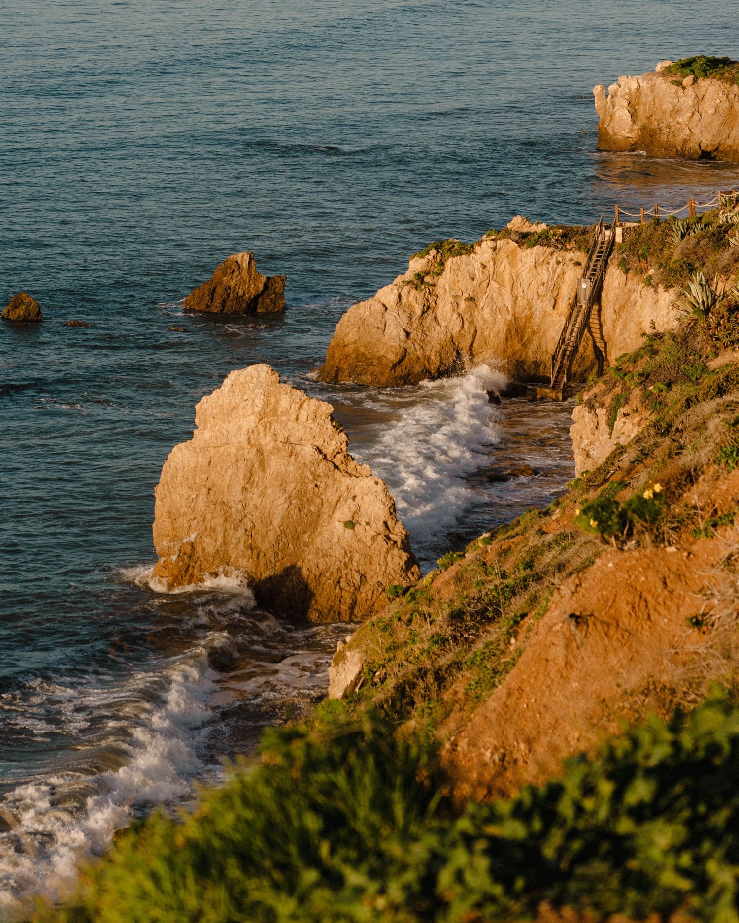 Golden hour sunlight illuminates rocky coastal cliffs with crashing waves and lush green vegetation along a scenic shoreline.