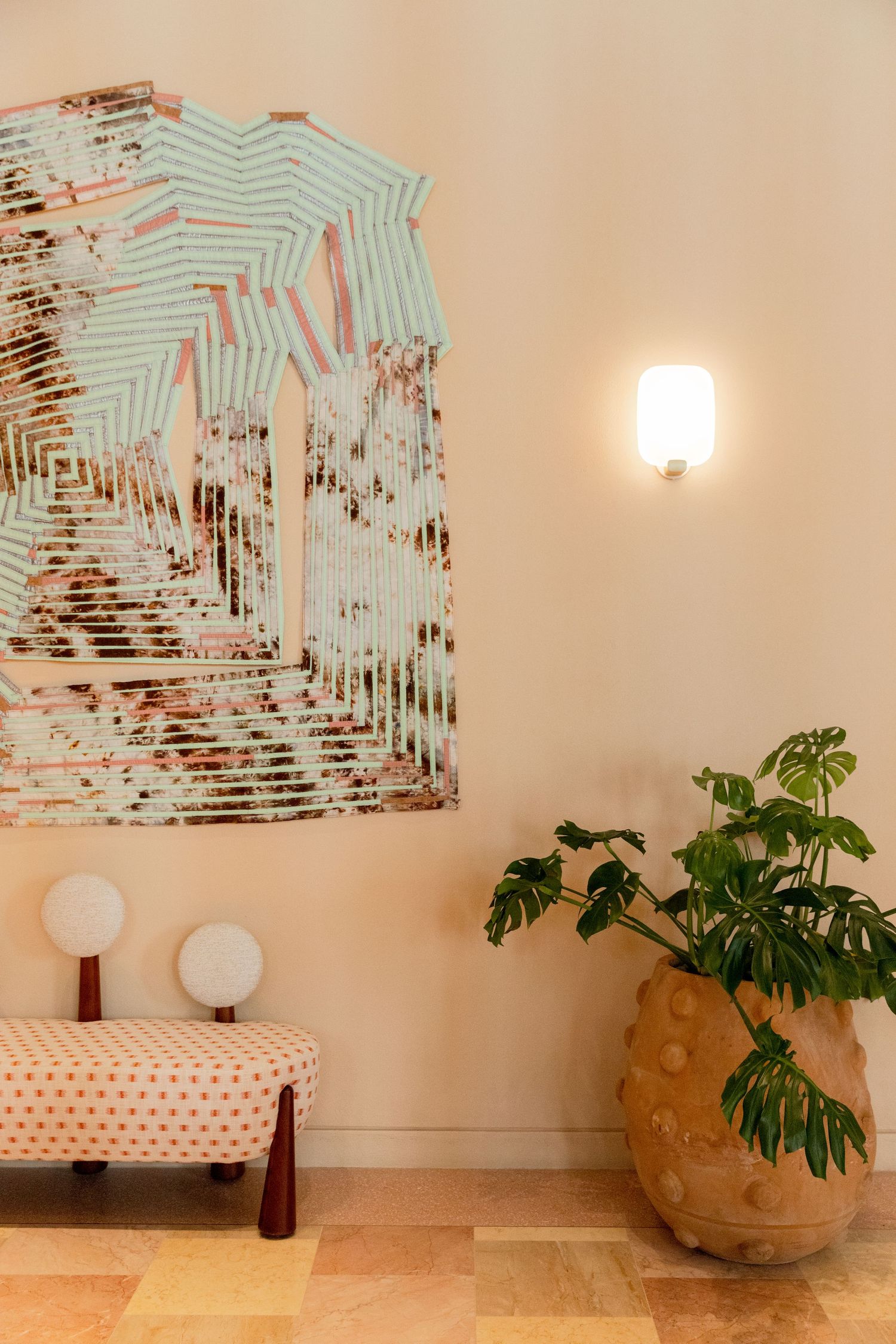 Pastel interior with abstract art print, polka dot bench, globe lamps, monstera plant in terracotta pot, and wall sconce.
