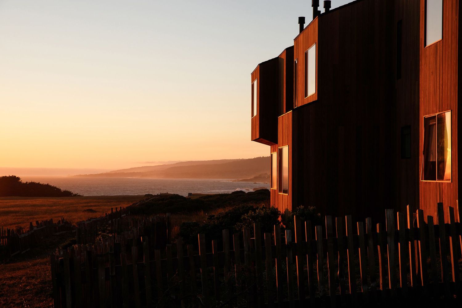 Modern wooden building with large windows at sunset, overlooking a scenic landscape with a rustic fence in the foreground.