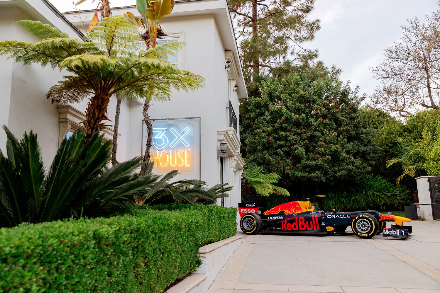 Red and black Formula 1 race car parked in front of a modern white house with manicured hedges.