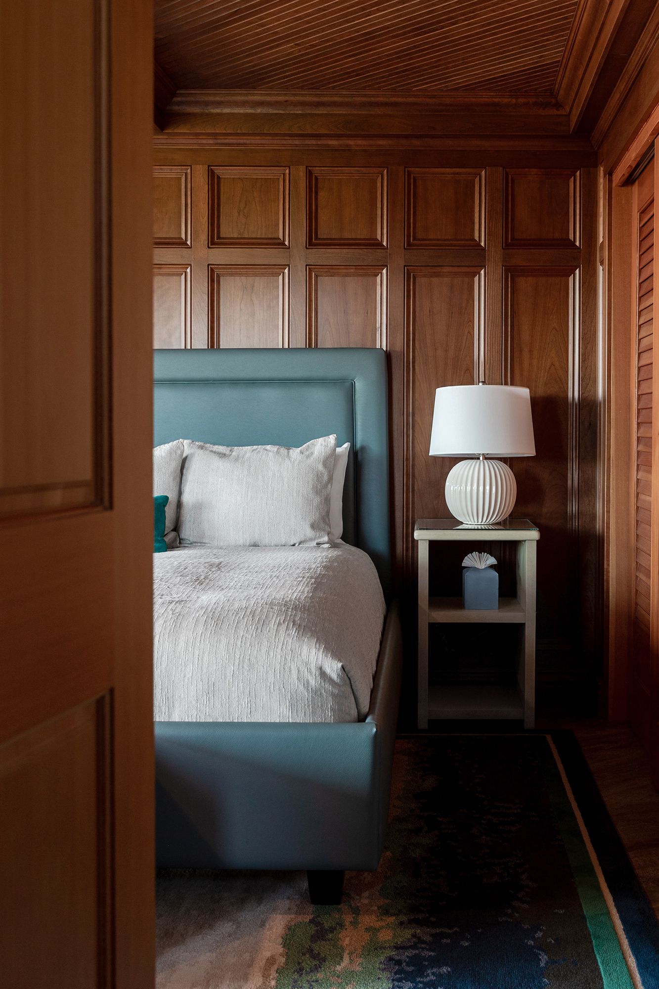Modern bedroom with teal upholstered bed frame, wood paneled walls, white bedding and ribbed ceramic table lamp on nightstand.