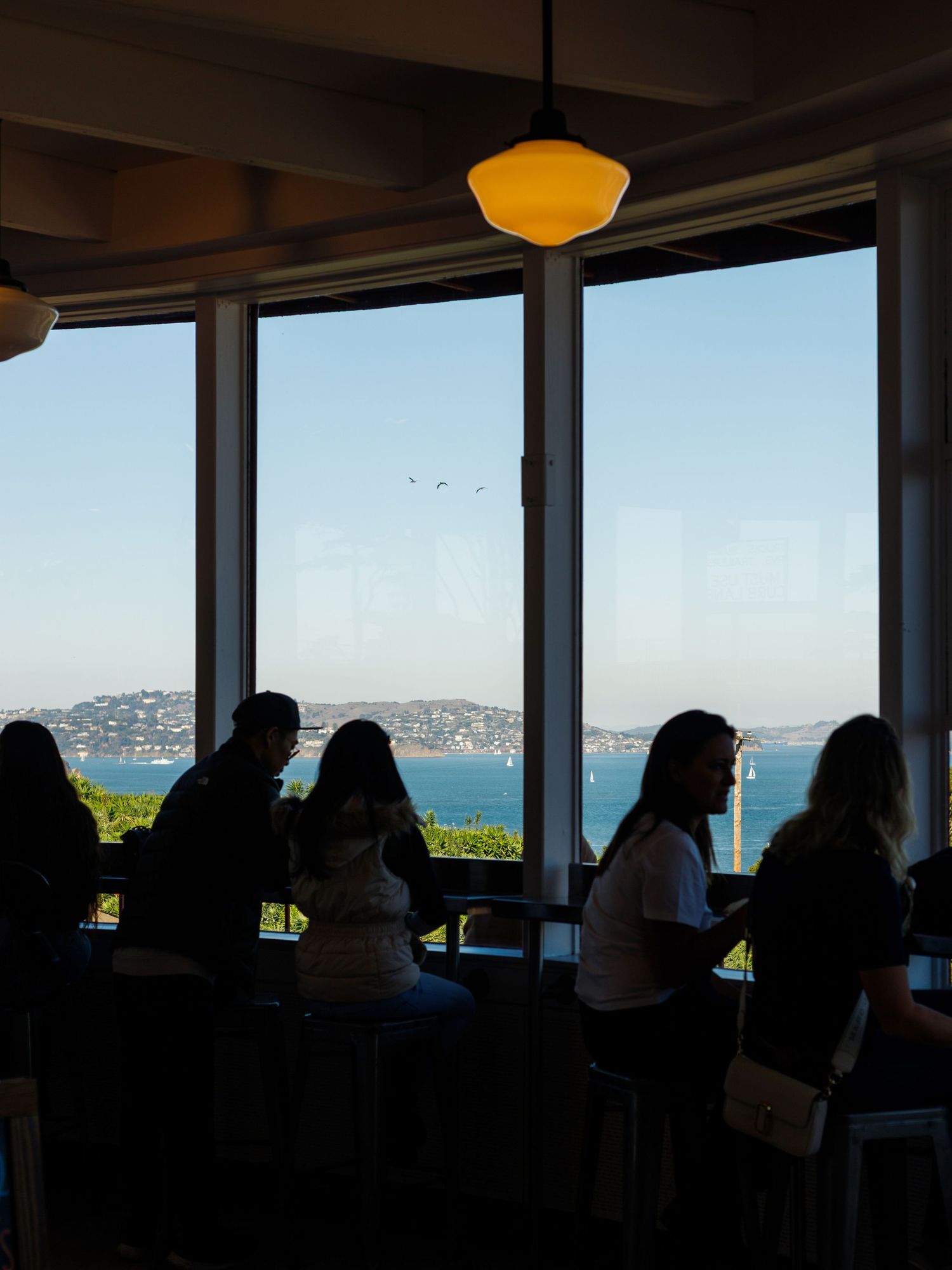Silhouetted people sit by large windows overlooking a scenic bay with blue skies in a restaurant.