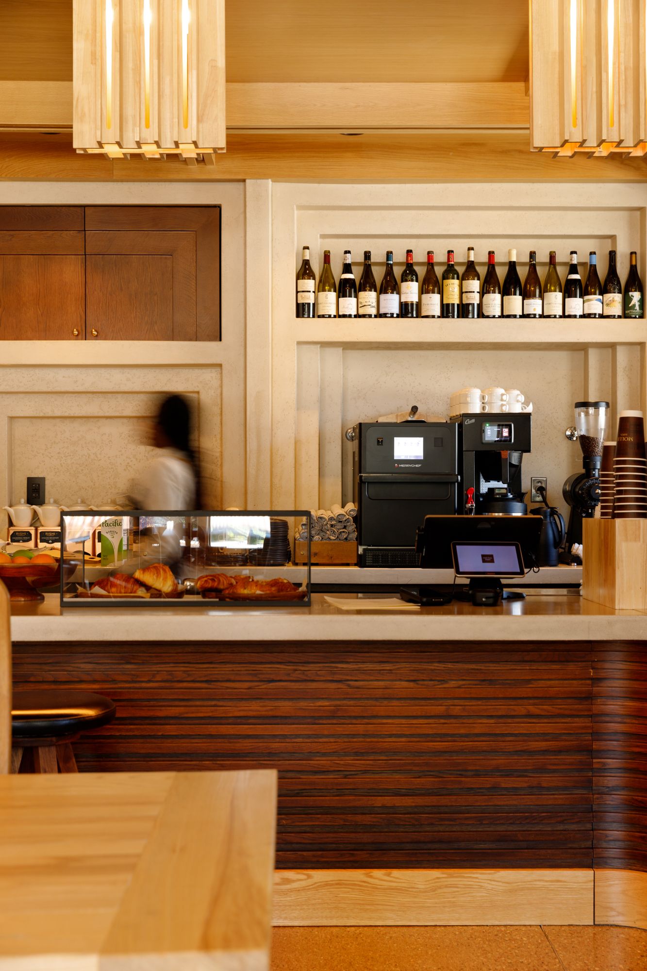 Warm wooden cafe bar counter with espresso machine, pastries, and wine bottles on shelves.