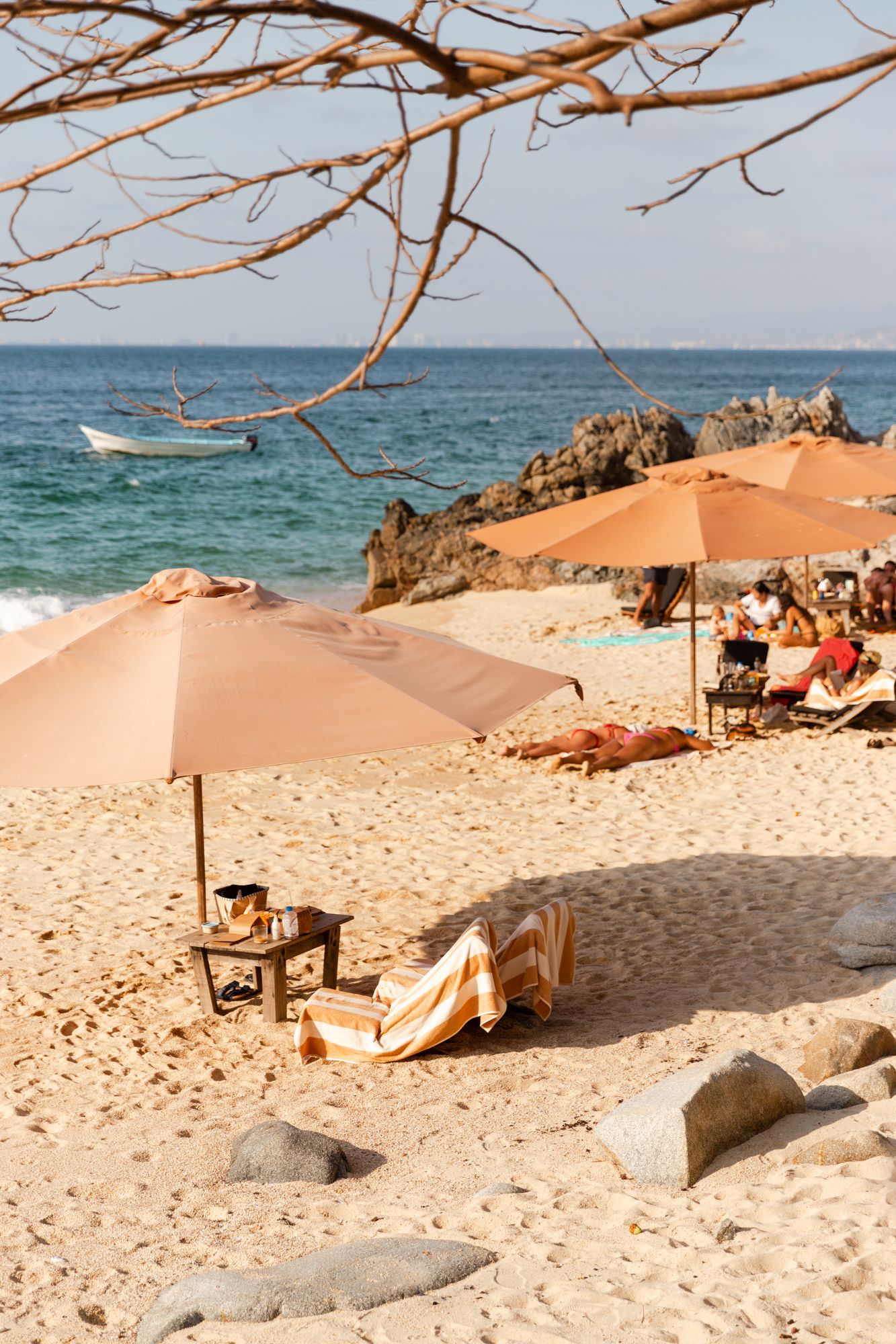 Orange beach umbrellas line a pristine sandy beach with turquoise ocean waters and rocky outcrops in the background.