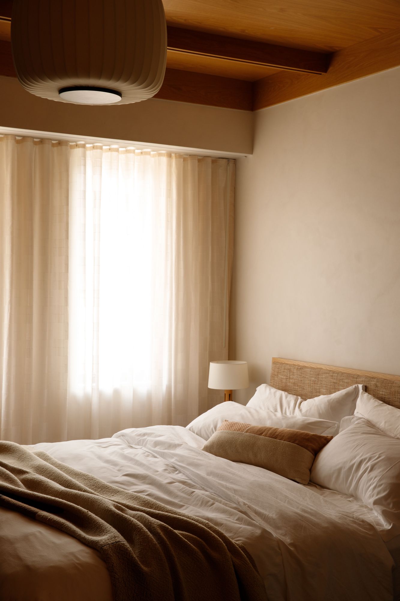 Serene hotel bedroom with white linen bedding, sheer curtains, rattan headboard, and warm pendant light.