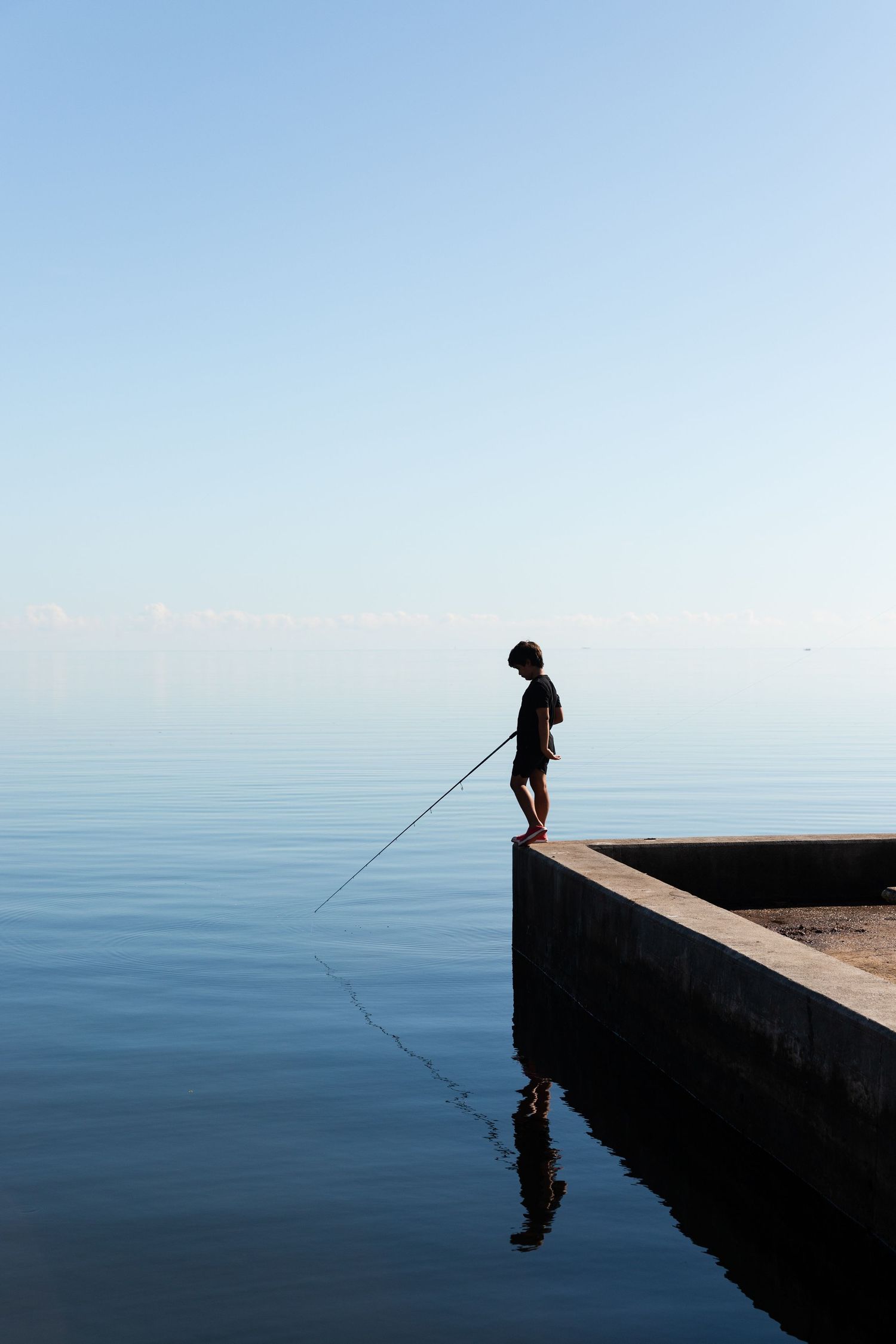 Silhouetted figures fishing from a concrete pier against a minimalist blue horizon.