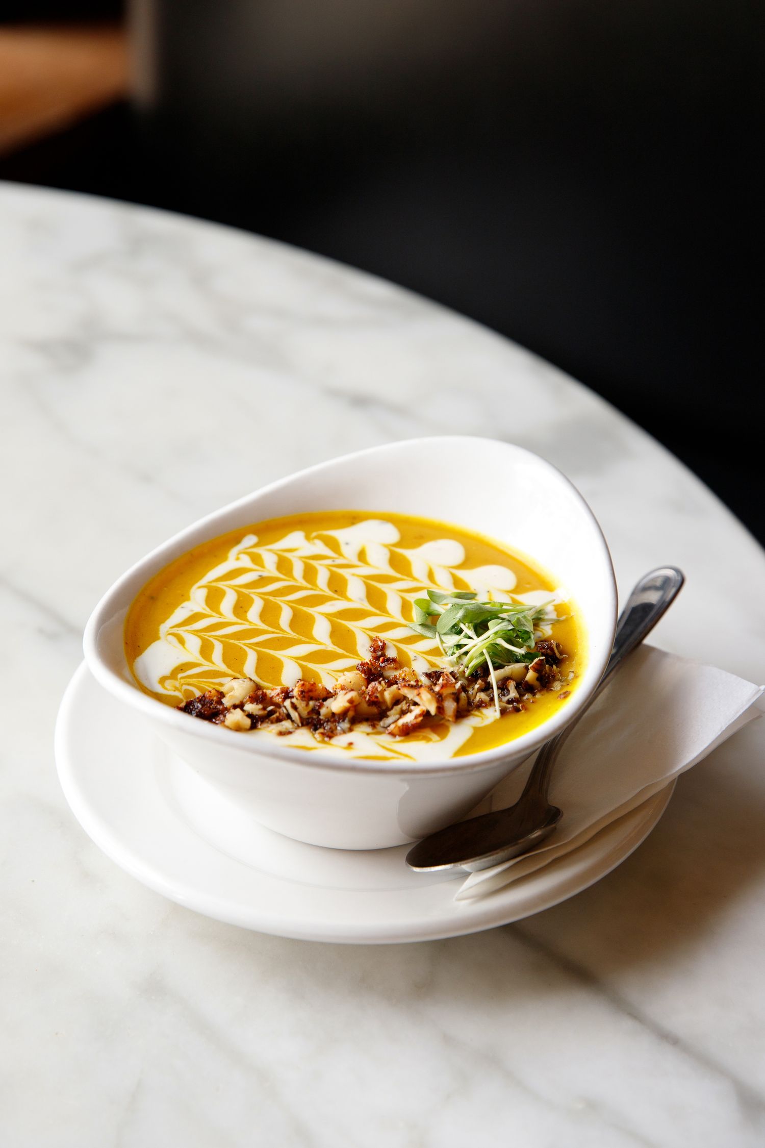 Creamy butternut squash soup with cream art, candied nuts, and microgreens in a white bowl on marble table.