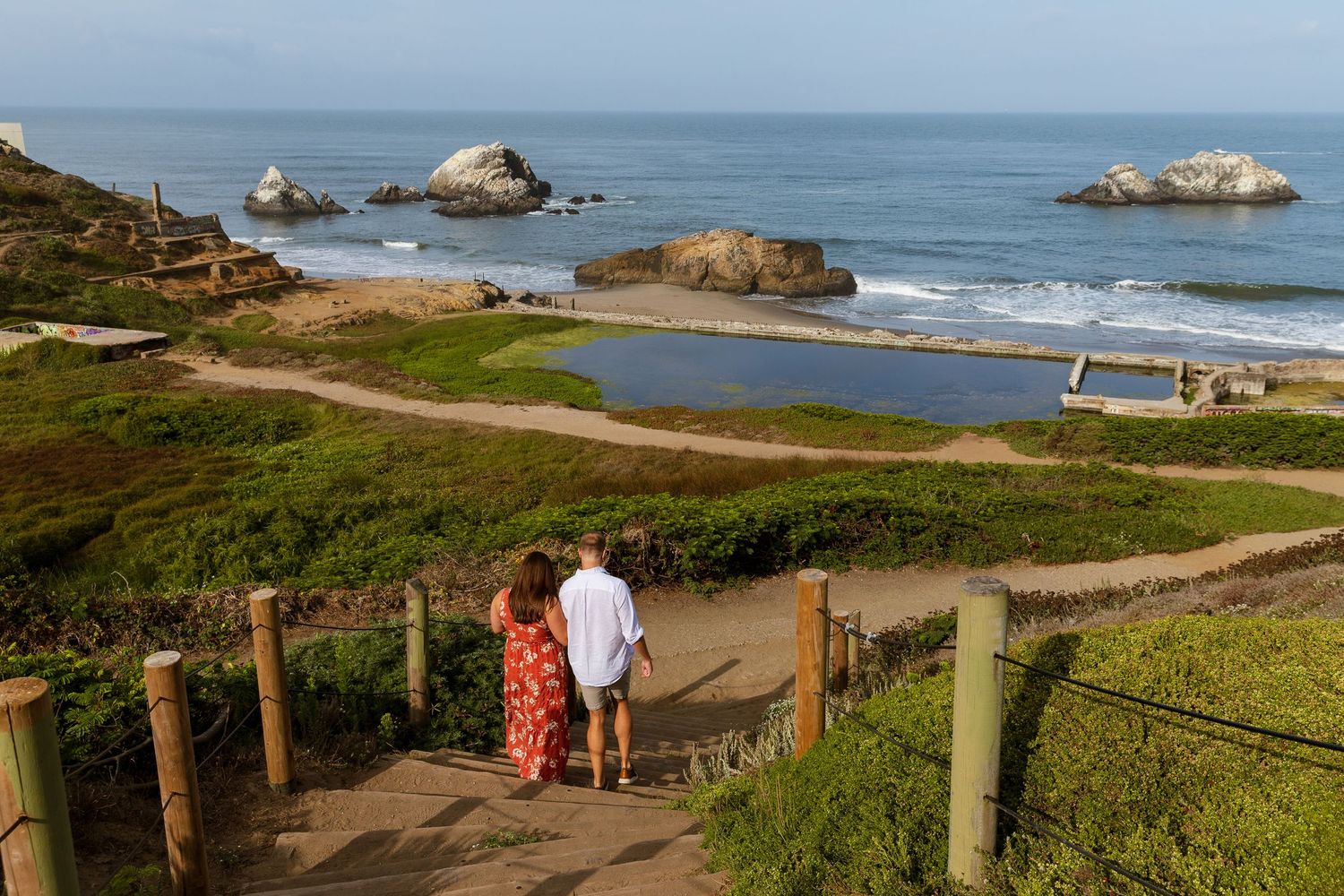 Couple walking down wooden steps at Sutro Baths ruins overlooking rocky coastline and Pacific Ocean in San Francisco.