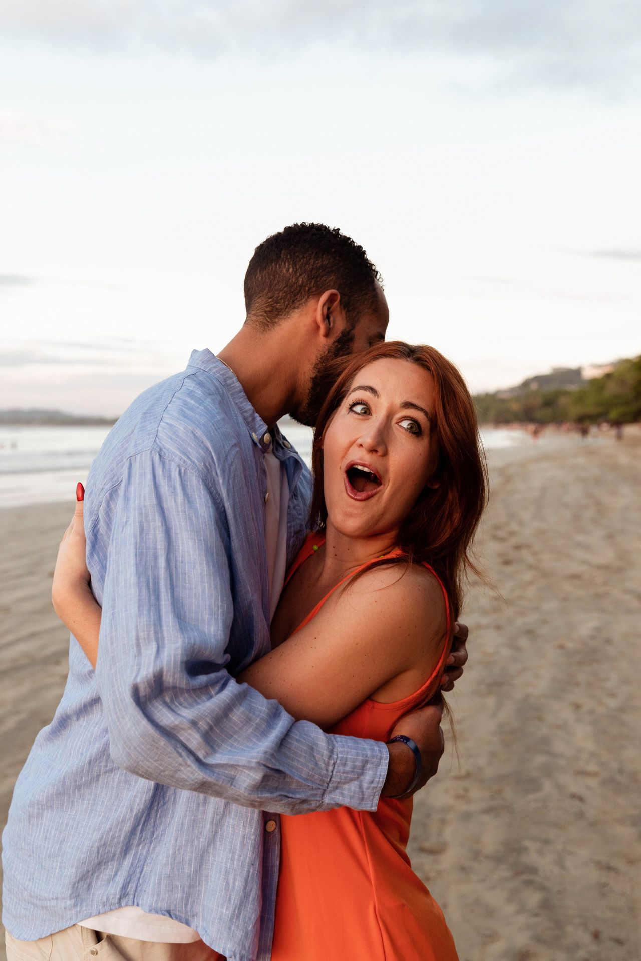 A couple shares a joyful moment embracing on a beach at sunset, with the woman wearing a coral dress.
