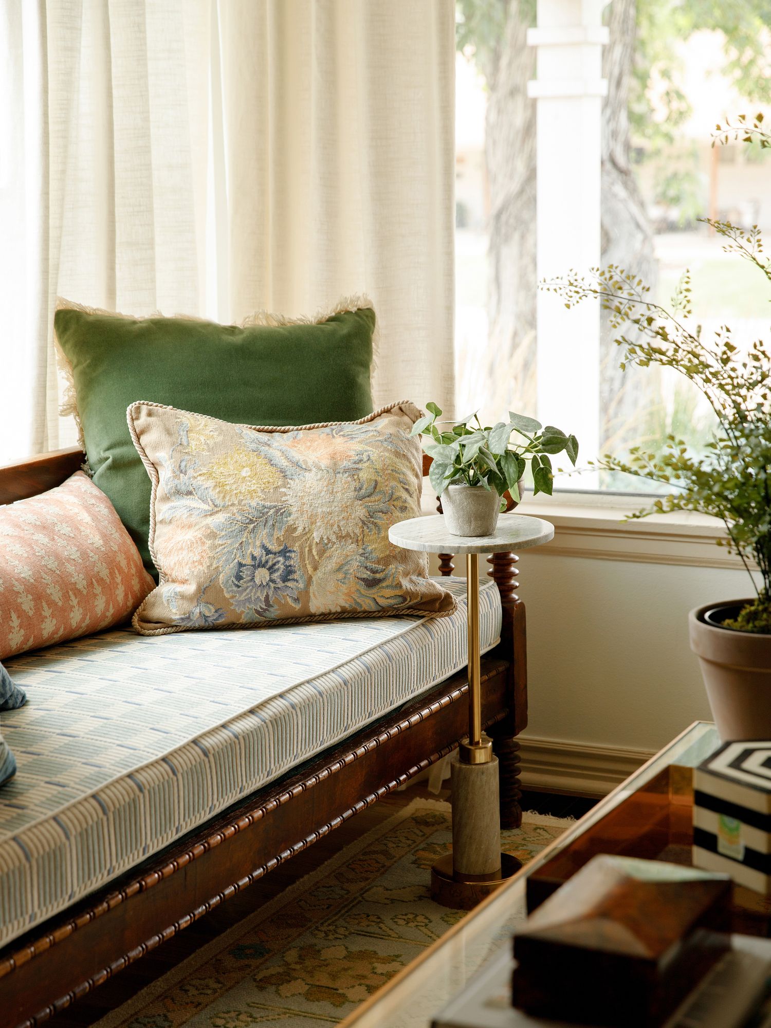 Elegant daybed with floral and green velvet pillows beside a small round side table with a plant near bright windows.