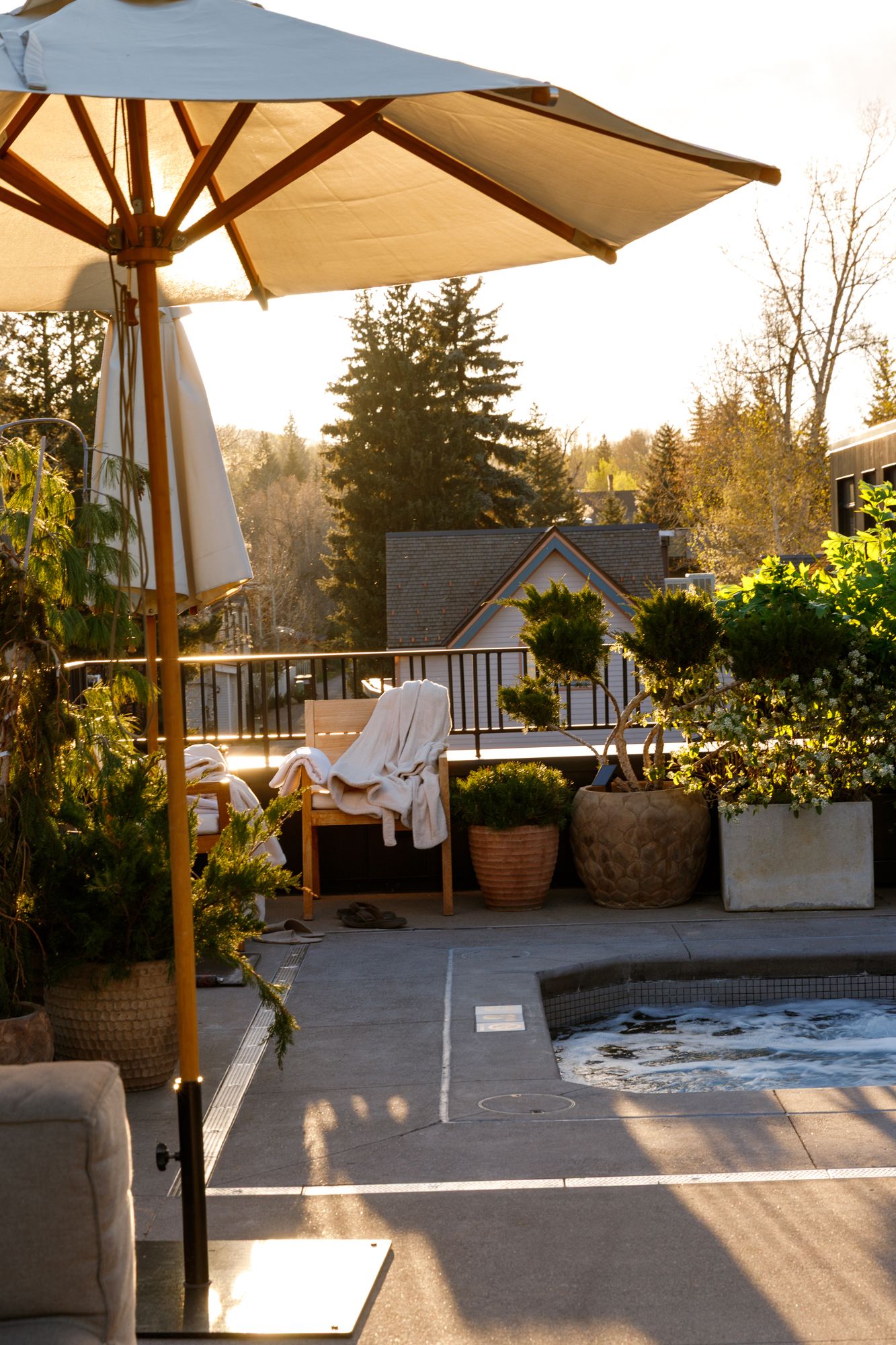 Sunny rooftop patio with umbrella, hot tub, and lush potted plants in warm golden light.