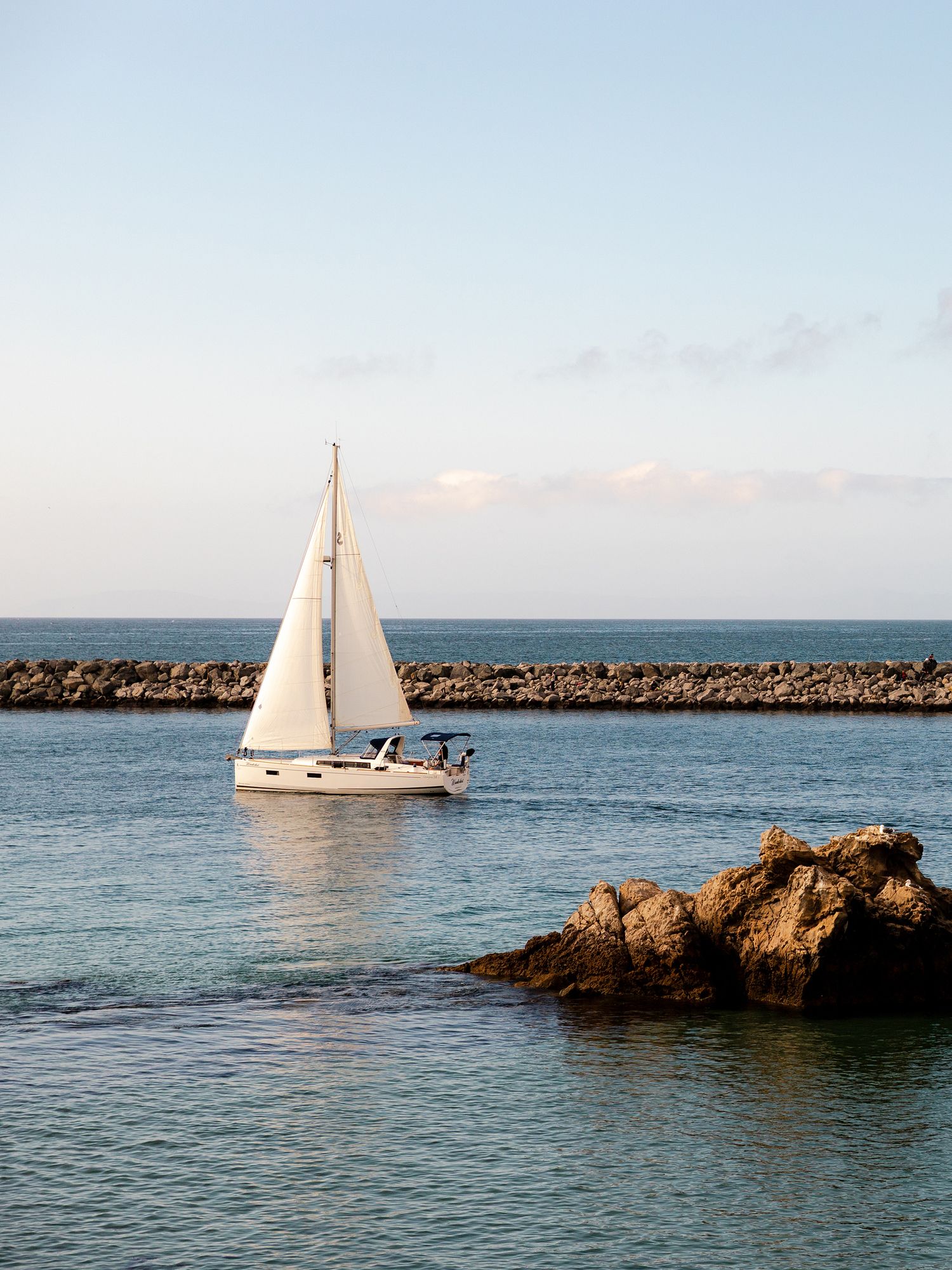 White sailboat gliding through calm blue waters near a rocky breakwater under a clear sky.