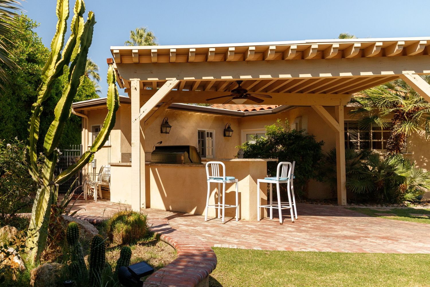 Outdoor patio with wooden pergola, outdoor kitchen, bar stools, cactus garden, and lush landscaping on a sunny day.