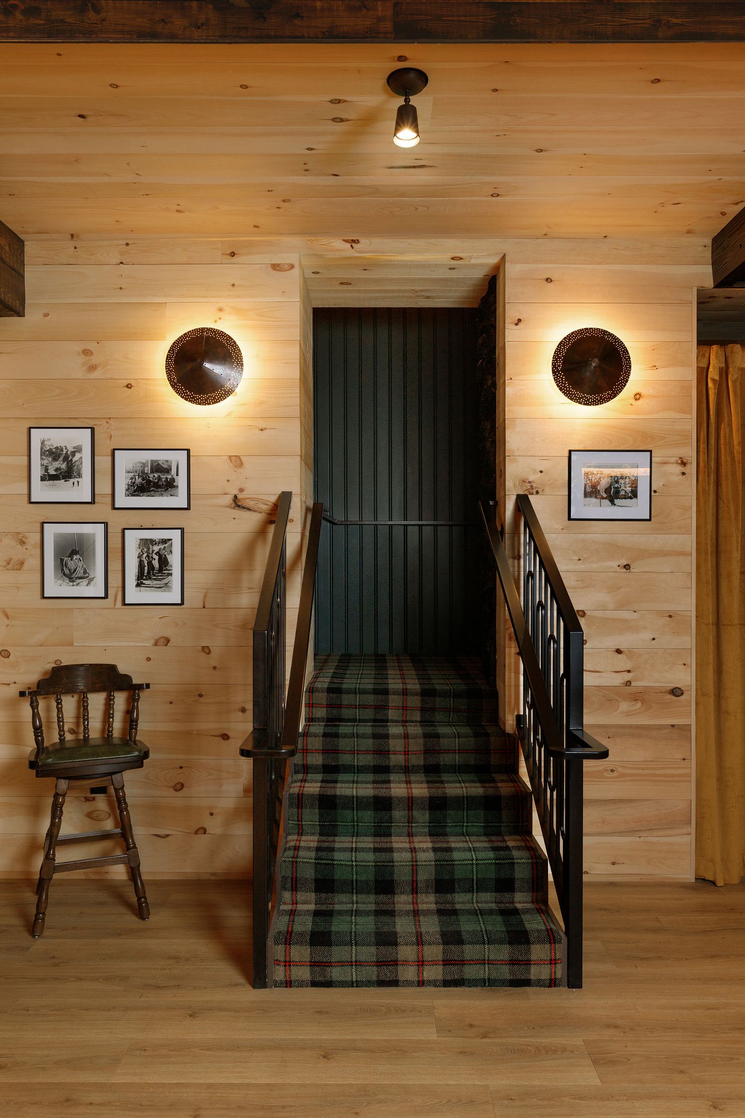 Rustic cabin staircase with plaid carpet runner, pine wood walls, wall sconces, framed art, and a vintage wooden chair.