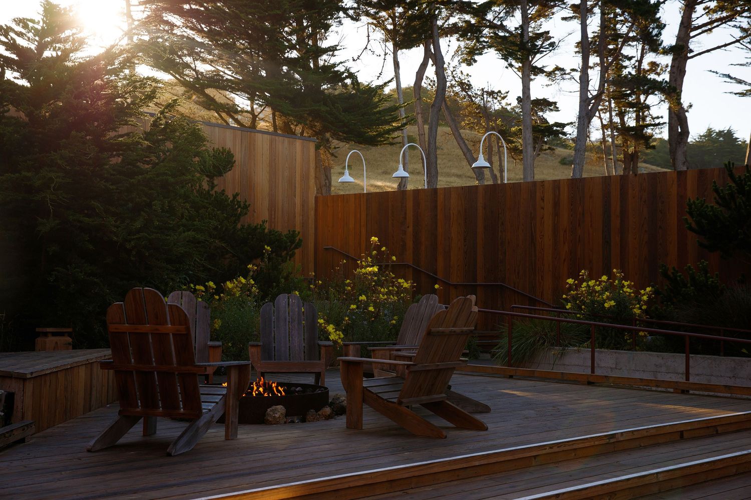 Wooden Adirondack chairs around a fire pit on a deck surrounded by trees and flowers at sunset.