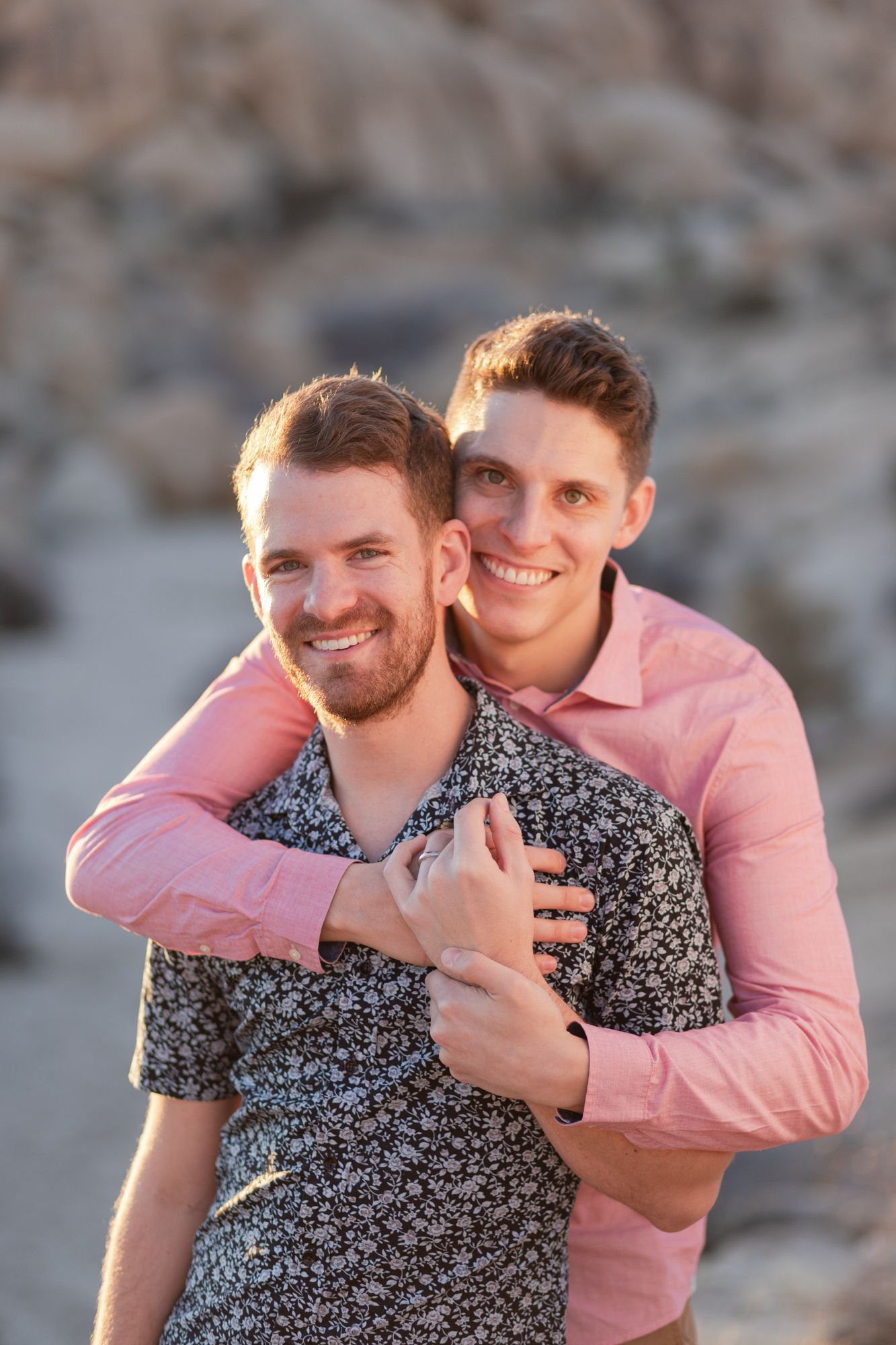A couple embraces outdoors while wearing pink and floral print shirts against a blurred natural background.