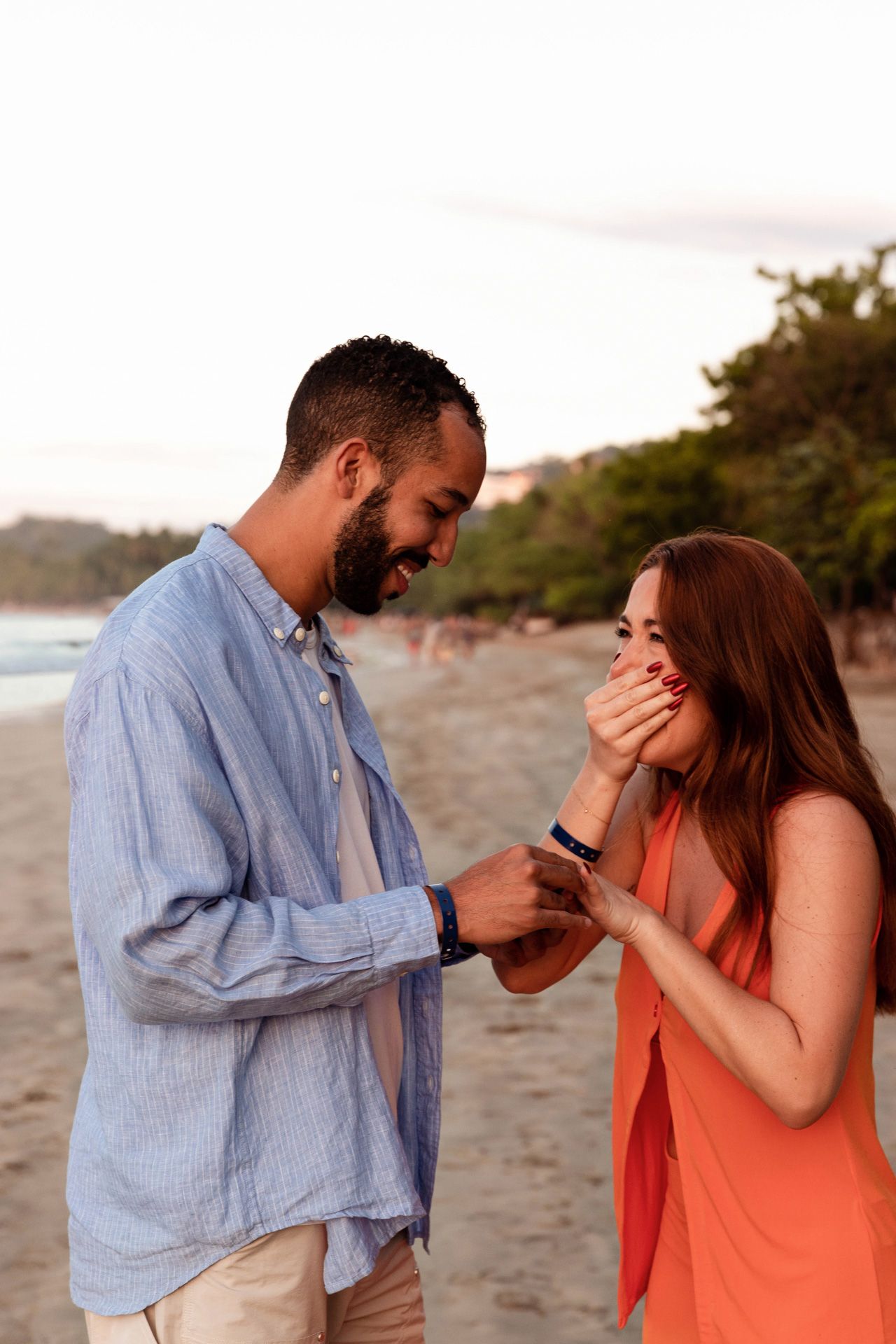 A couple shares an emotional moment during a beach proposal at sunset, with her surprised reaction captured.
