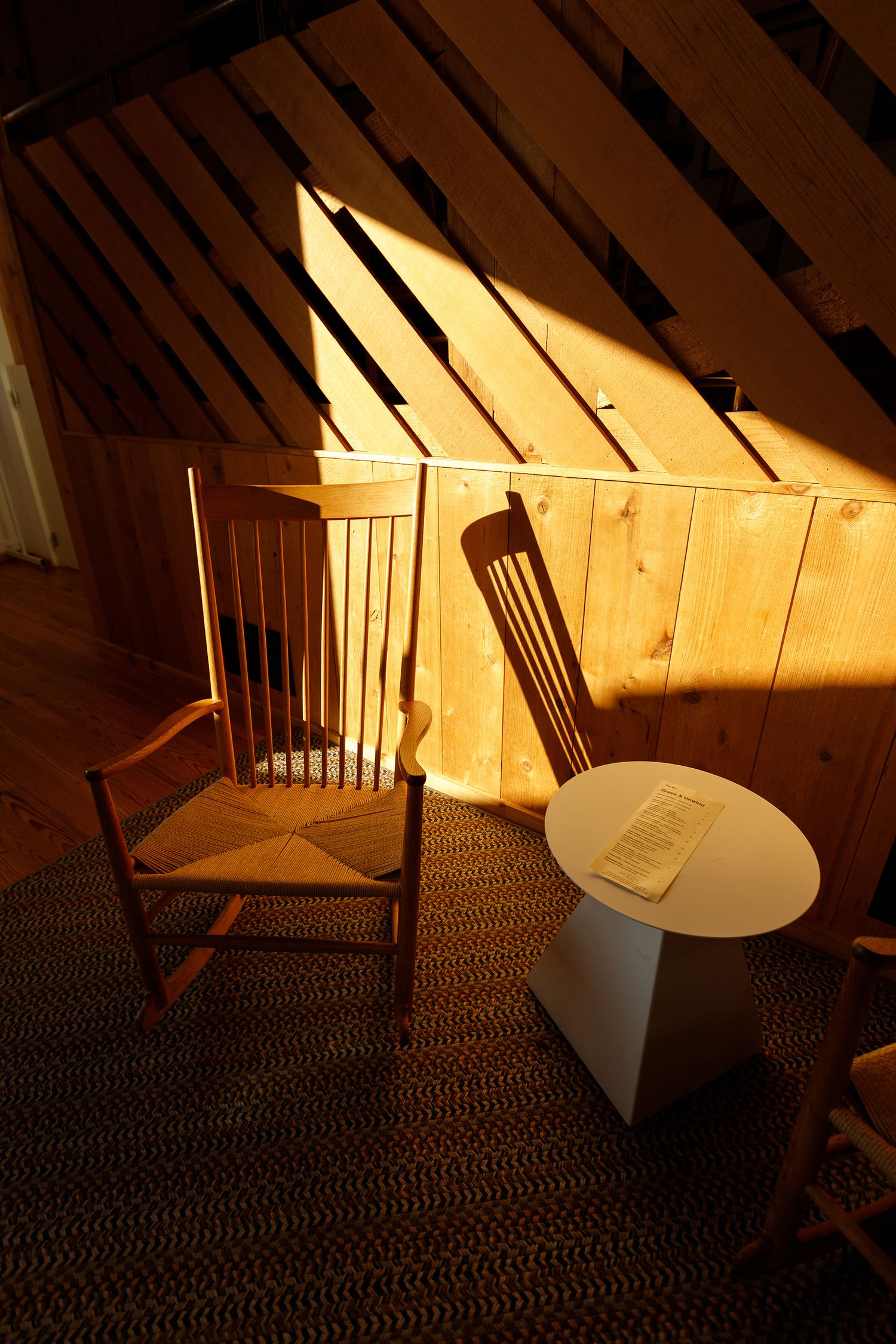 Rocking chair and side table bathed in warm golden light inside a rustic wood-paneled attic room.