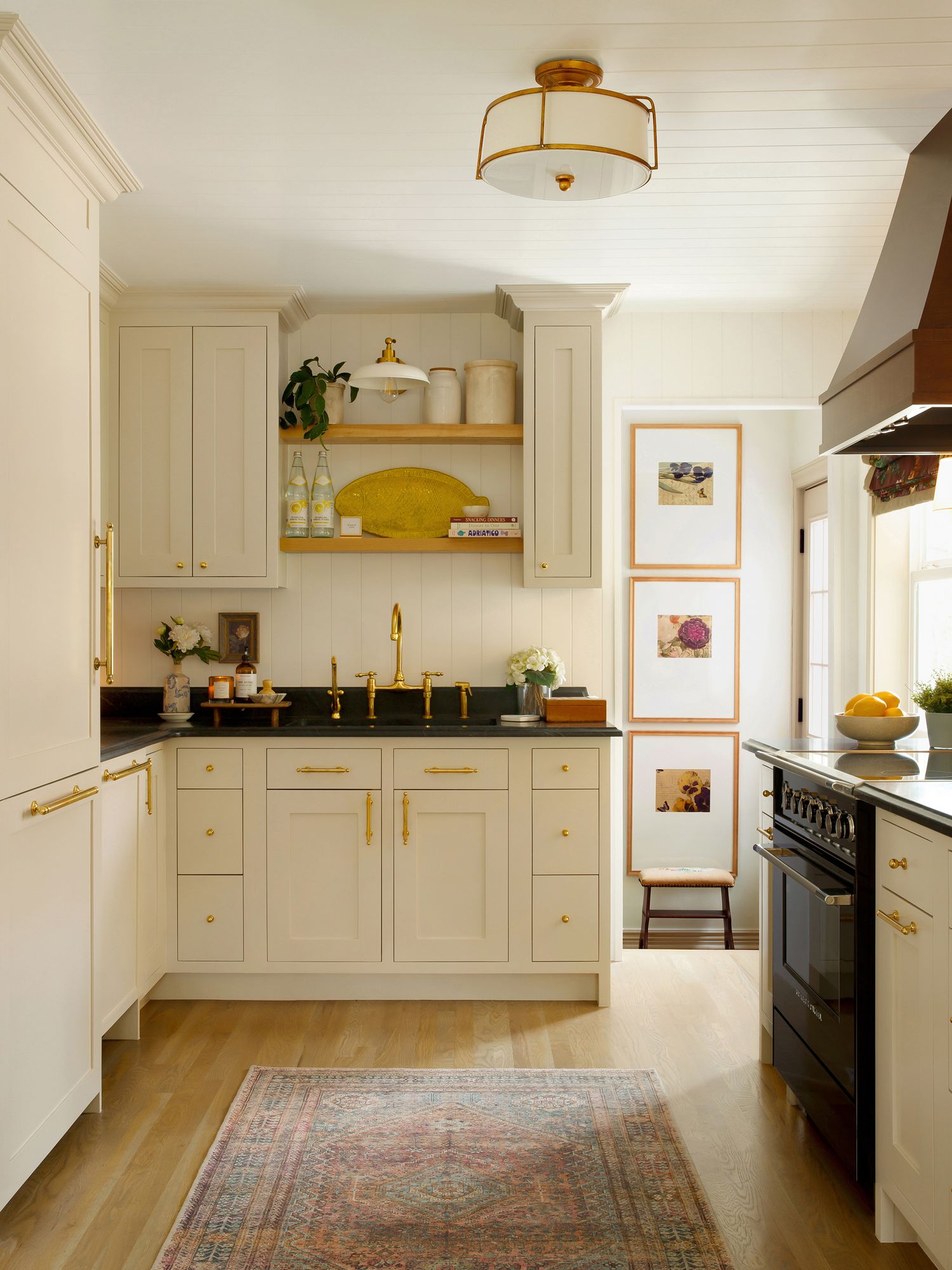 Bright kitchen with cream shaker cabinets, black countertops, brass hardware, open shelves, vintage rug, and framed art prints.