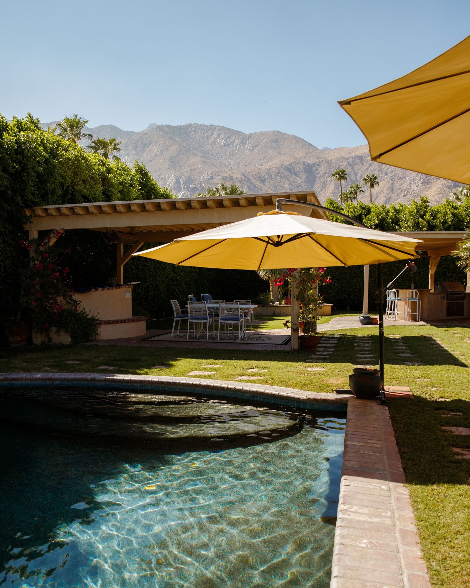 Outdoor pool area with yellow patio umbrellas, dining set, lush lawn, and mountain views at a desert resort.