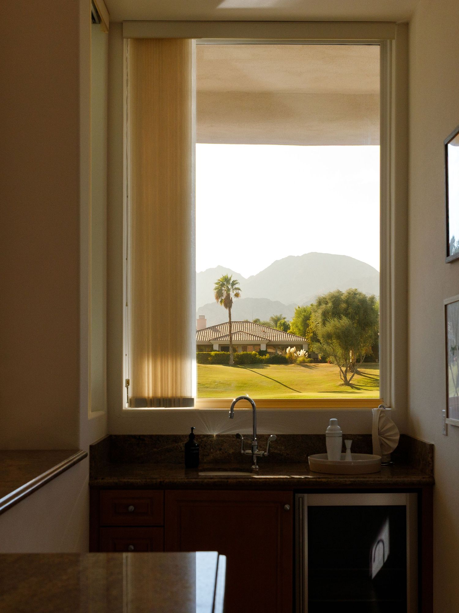 Wet bar with dark granite countertop and sink overlooking a scenic golf course with palm trees and mountains through a large window.