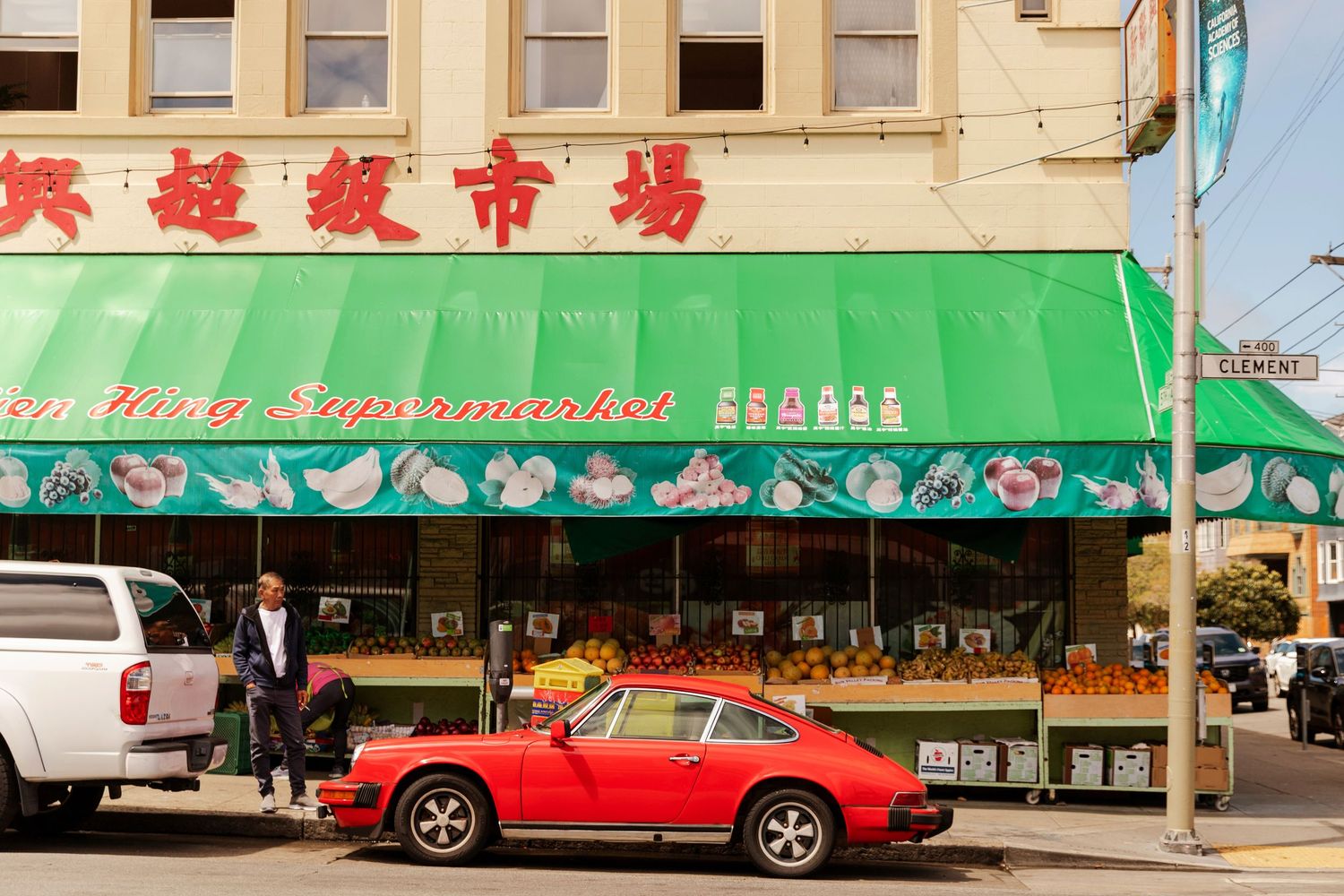 Red Porsche 911 parked outside a green-awning Asian supermarket in a vibrant urban Chinatown neighborhood.