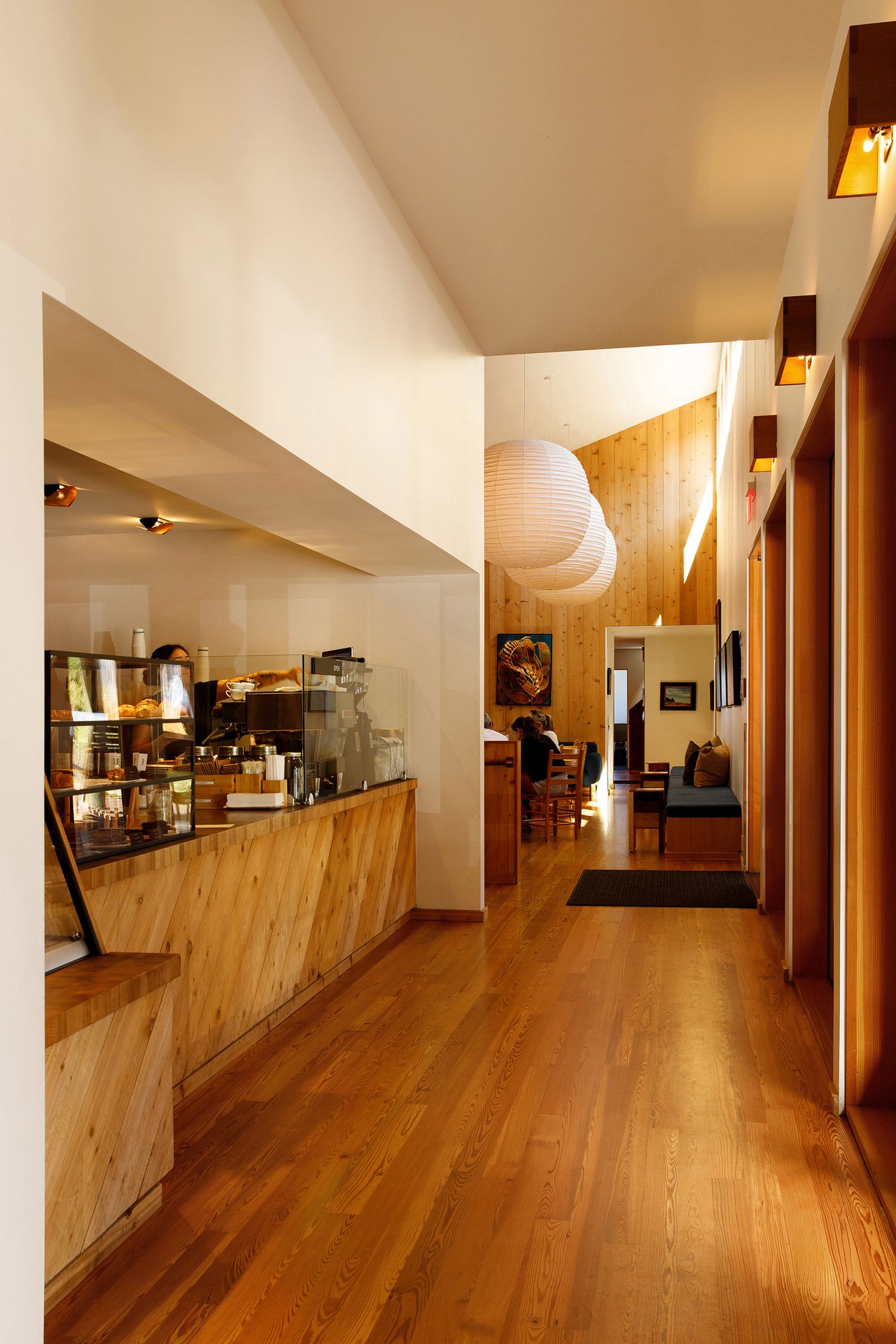 Modern cafe interior with warm wood floors, a service counter, pendant lighting, and an open dining area in the background.