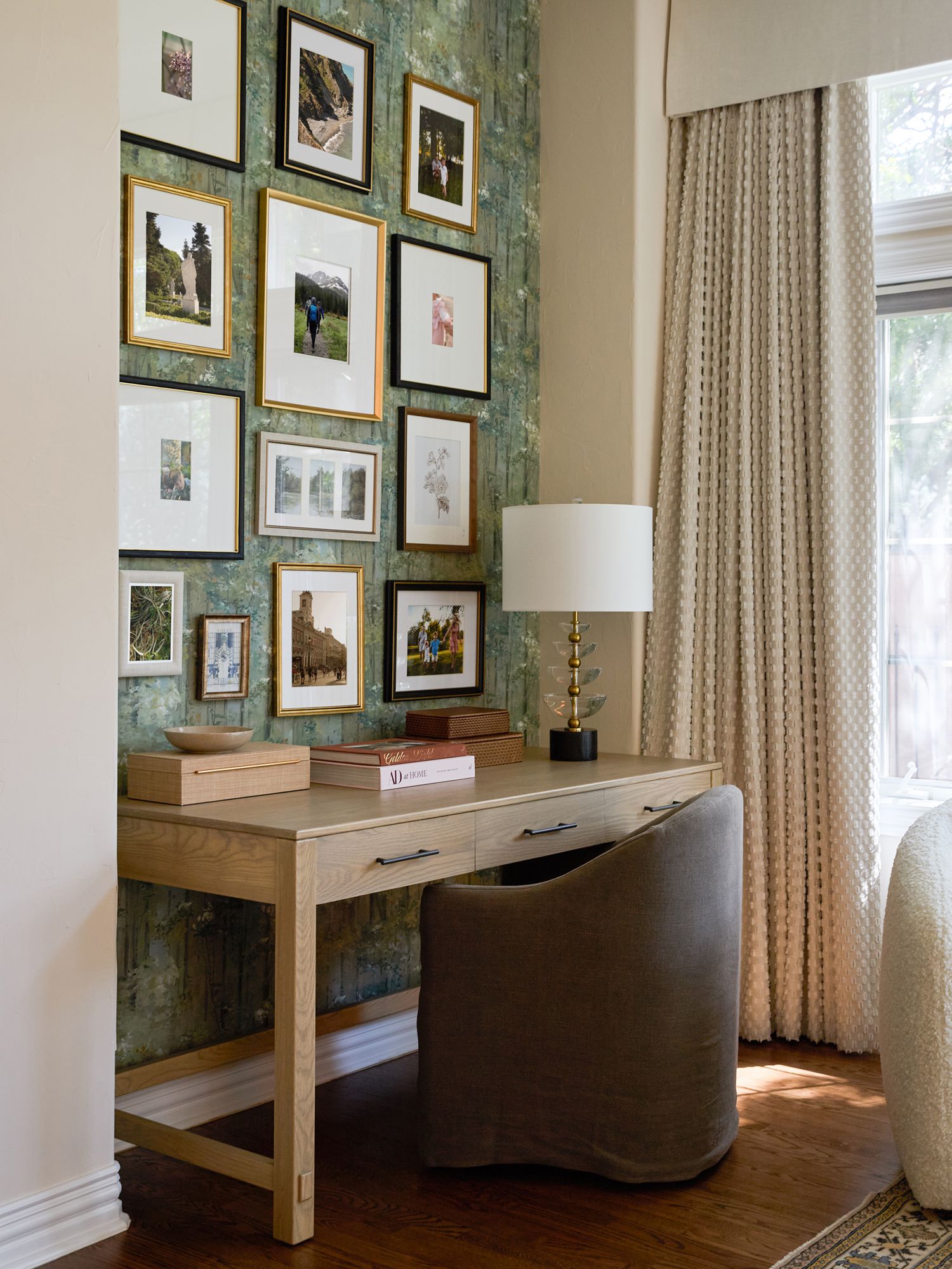 Light wood writing desk with a gray upholstered chair, brass base lamp, and a gallery wall of framed art on textured green wallpaper.