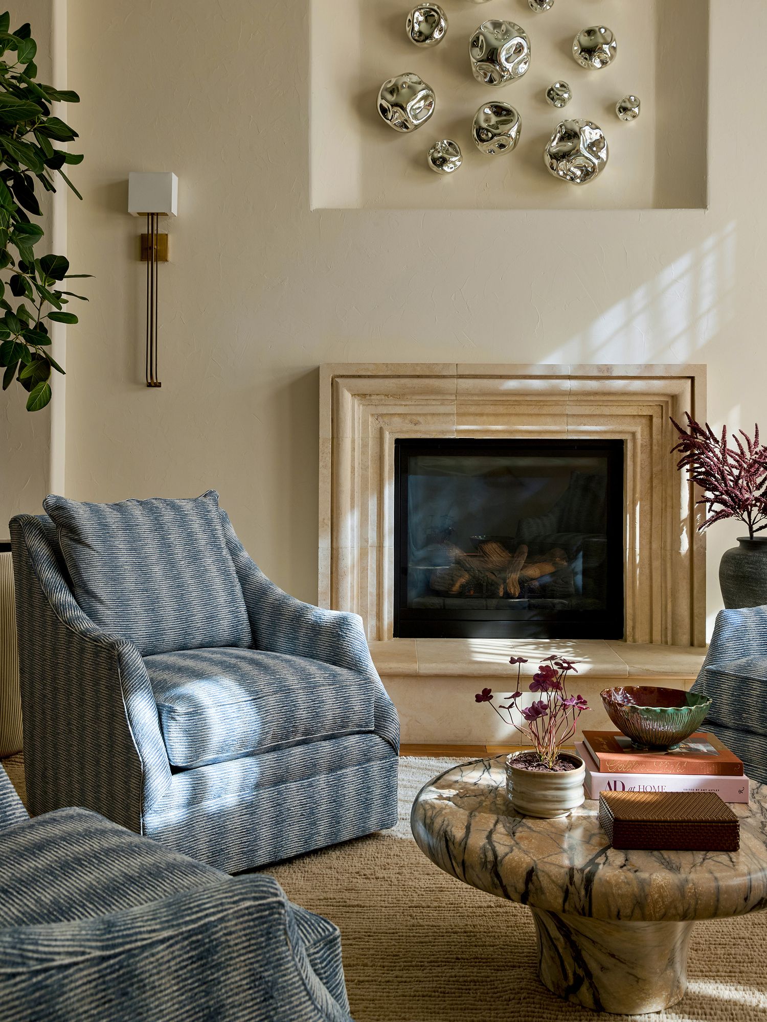 Elegant living room with blue striped armchairs, marble coffee table, stone fireplace, and silver wall art accents.