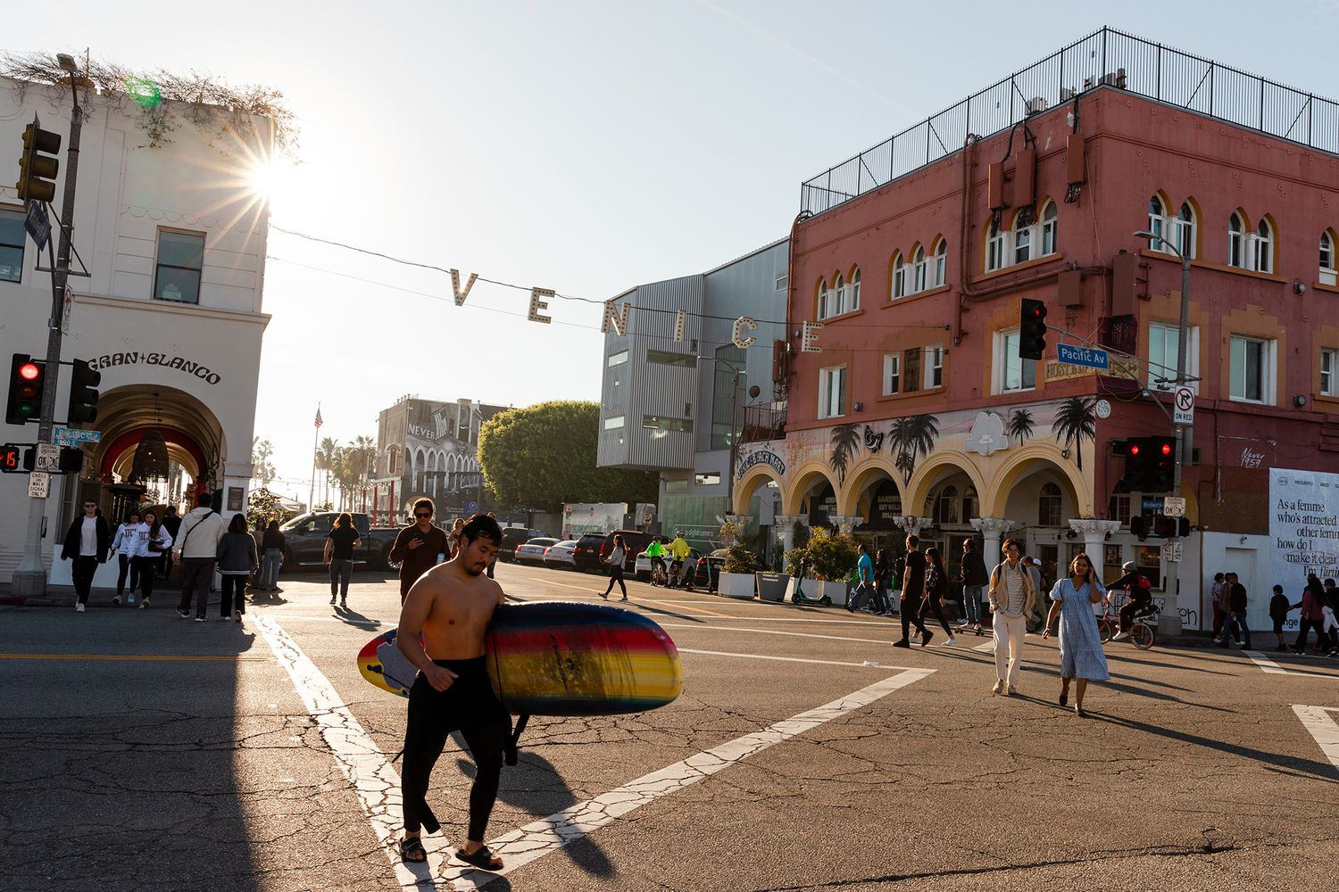 Sunny Venice Beach street scene with surfer carrying colorful board near iconic Venice sign and red buildings.