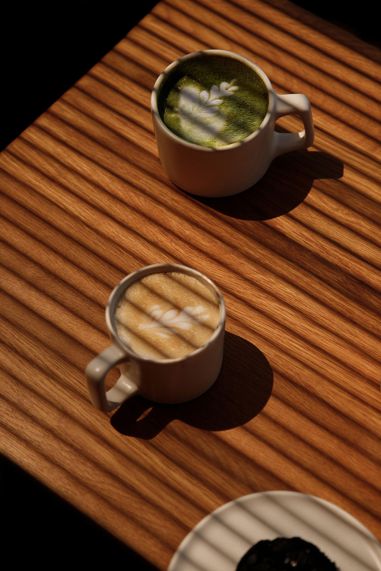 Two artisan lattes, one matcha and one classic, served in white ceramic mugs on a wooden table in warm sunlight.