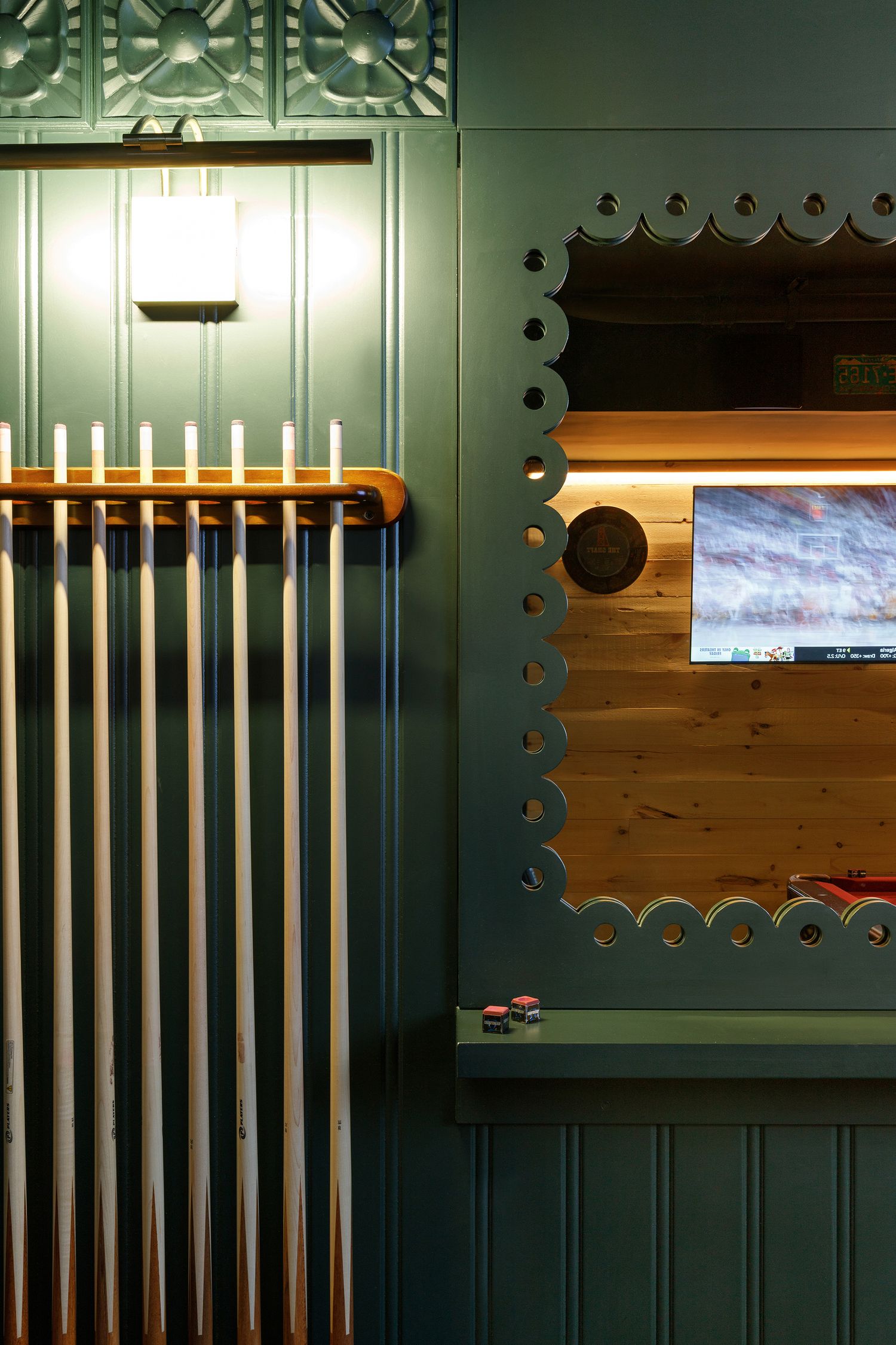 Green-paneled interior with decorative scalloped trim, wall-mounted TV, billiard cue rack, and warm accent lighting.