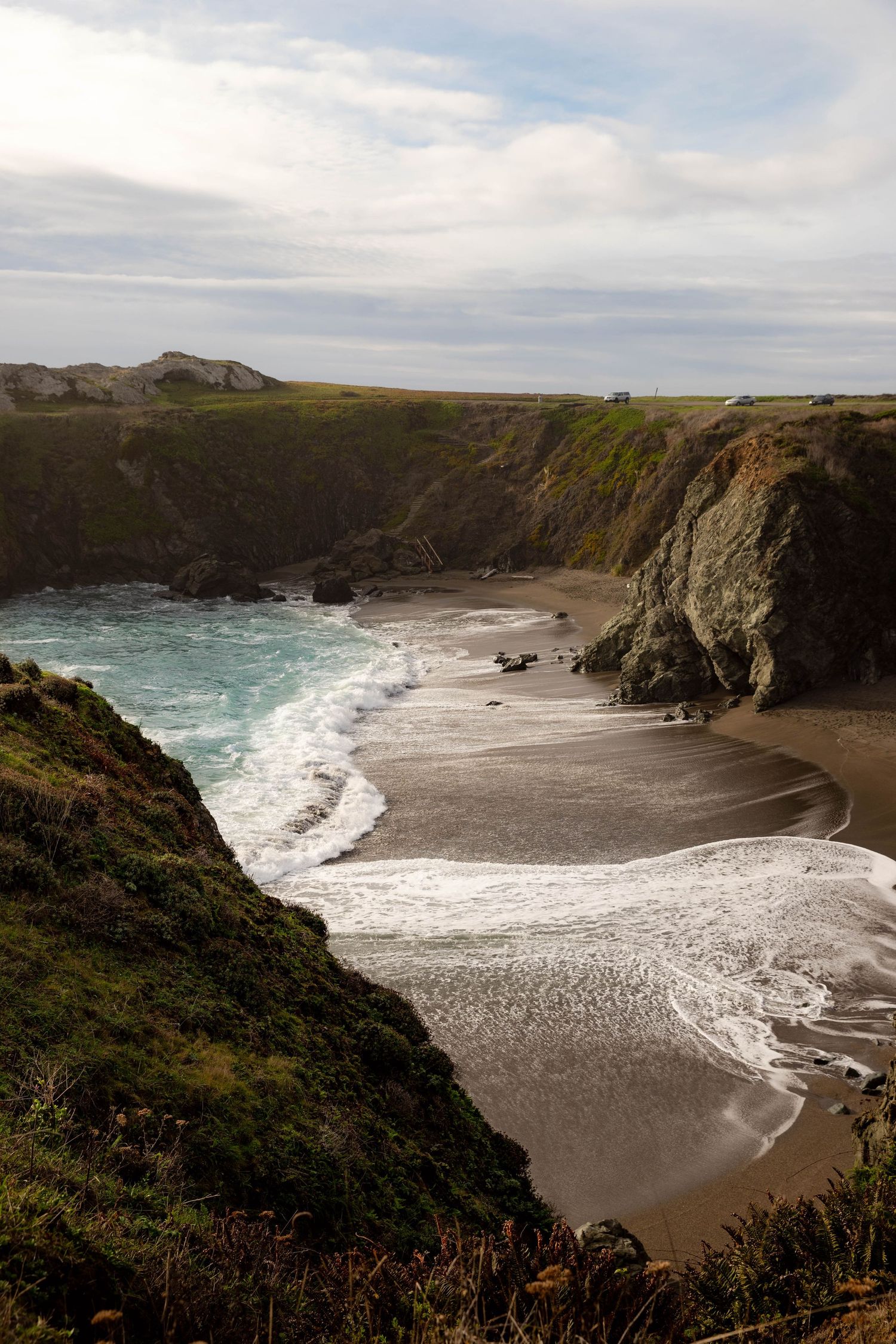 Dramatic coastal cliffs frame a secluded beach with crashing turquoise waves along a rugged shoreline under cloudy skies.