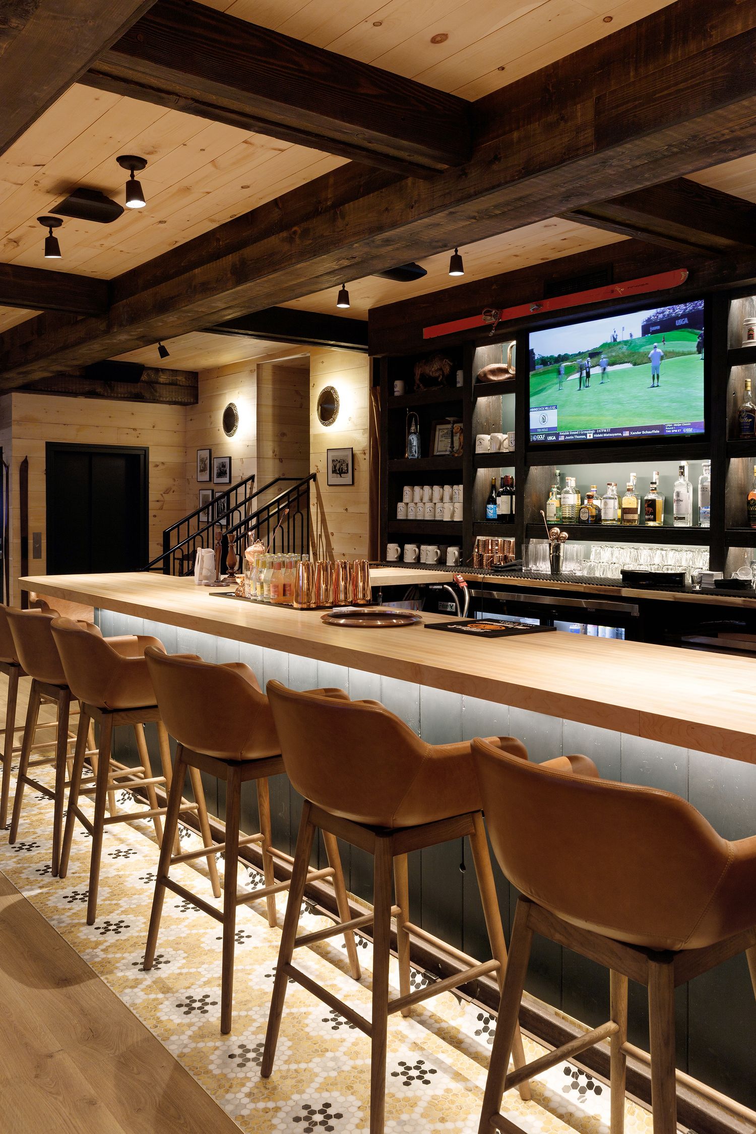 Elegant bar with leather stools, wood beam ceiling, bottle display shelves, and patterned tile floor.