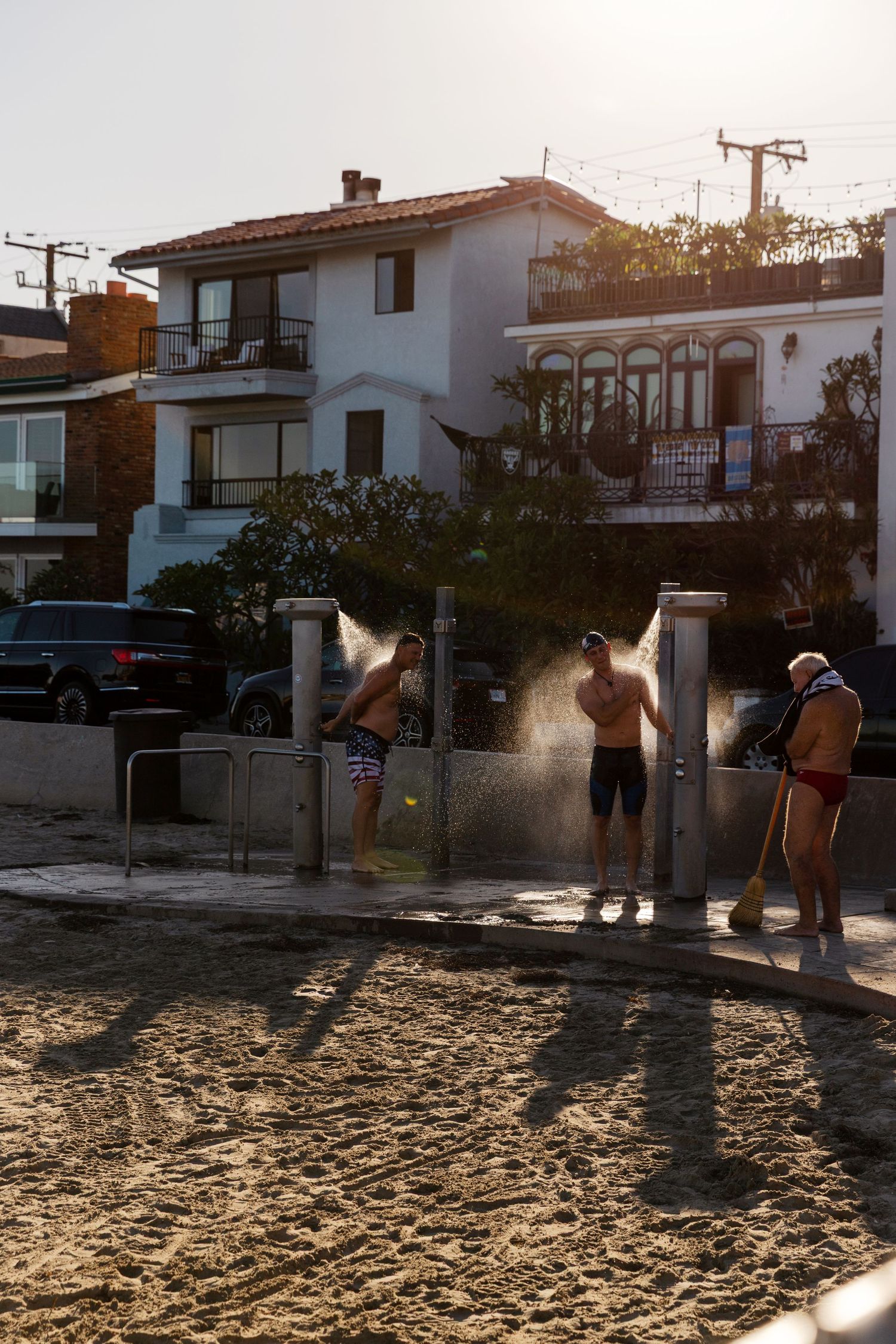 Three men rinse off at outdoor beach showers on sandy shore with coastal houses in background at golden hour.