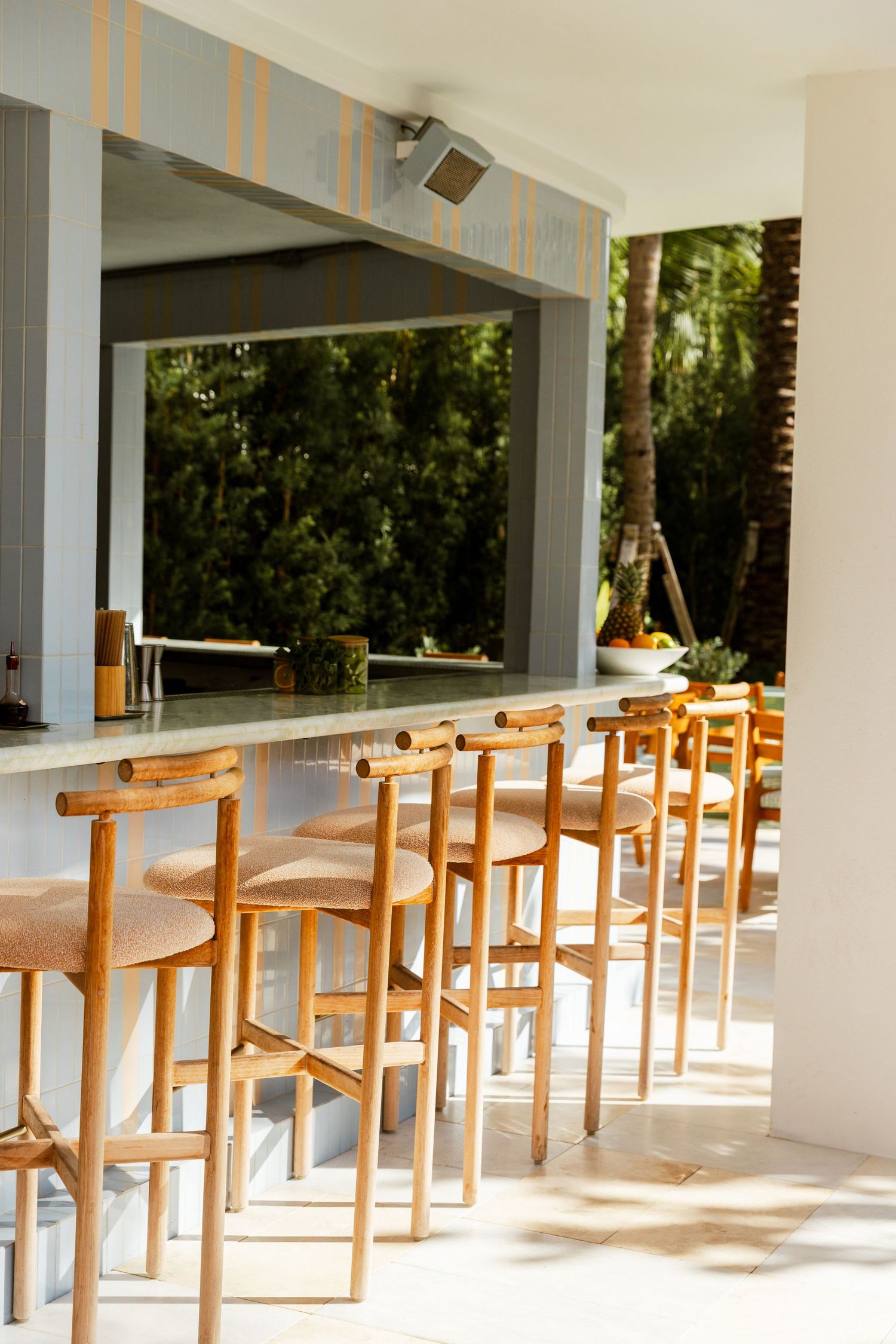 Outdoor bar area with wooden stools lined along a marble counter, open to lush greenery beyond.