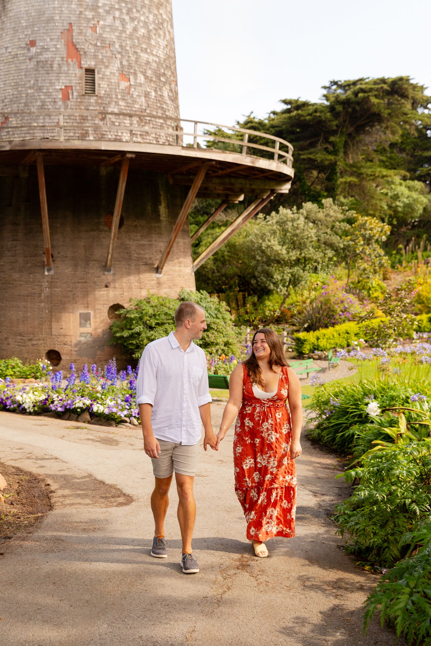 A couple walks hand in hand along a garden path near a lighthouse on a sunny summer day.
