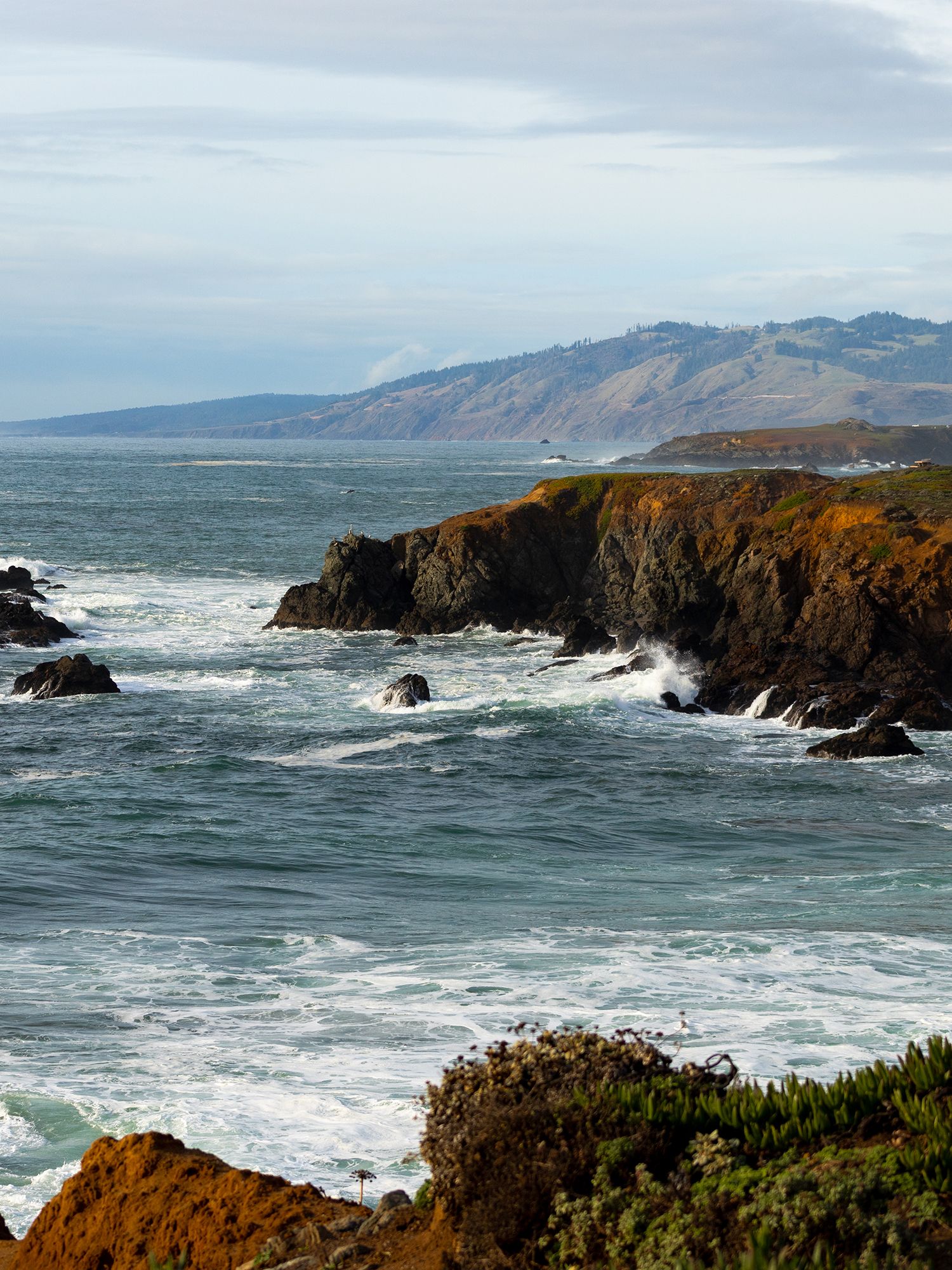 Rocky coastal cliffs with crashing waves and a misty mountain peninsula along the rugged California coastline.