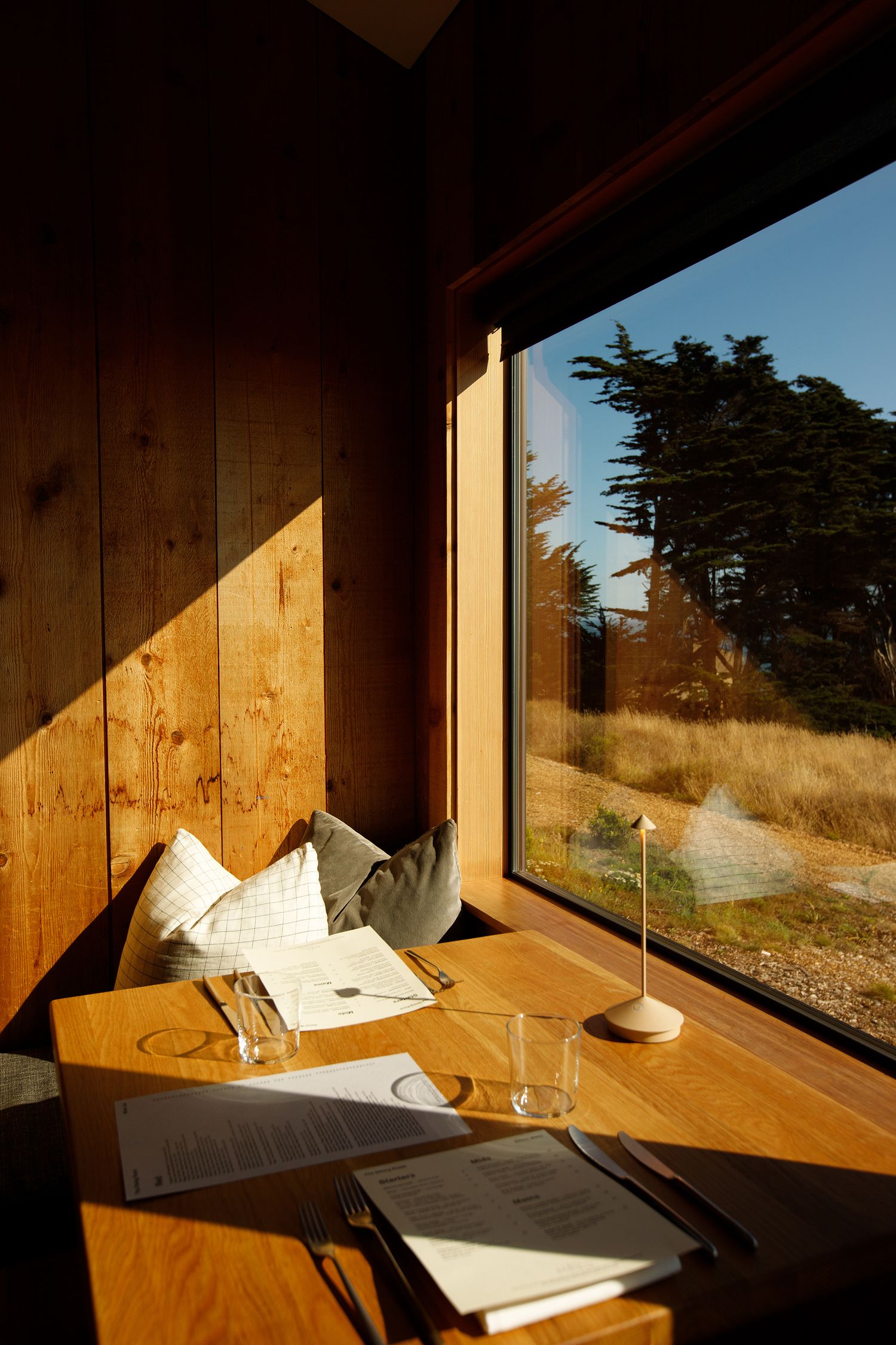 Cozy wooden booth dining nook with cushions, menus, and a small lamp beside a large window overlooking nature.