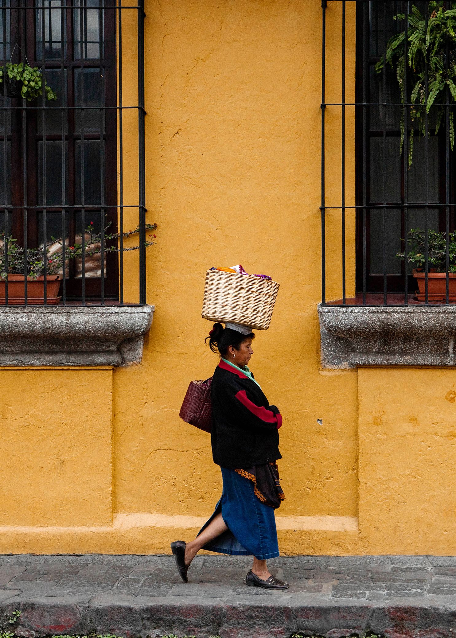 Guatemalan woman carrying baskets walks along a vibrant yellow wall with black-barred windows in sequence taken by travel photographer