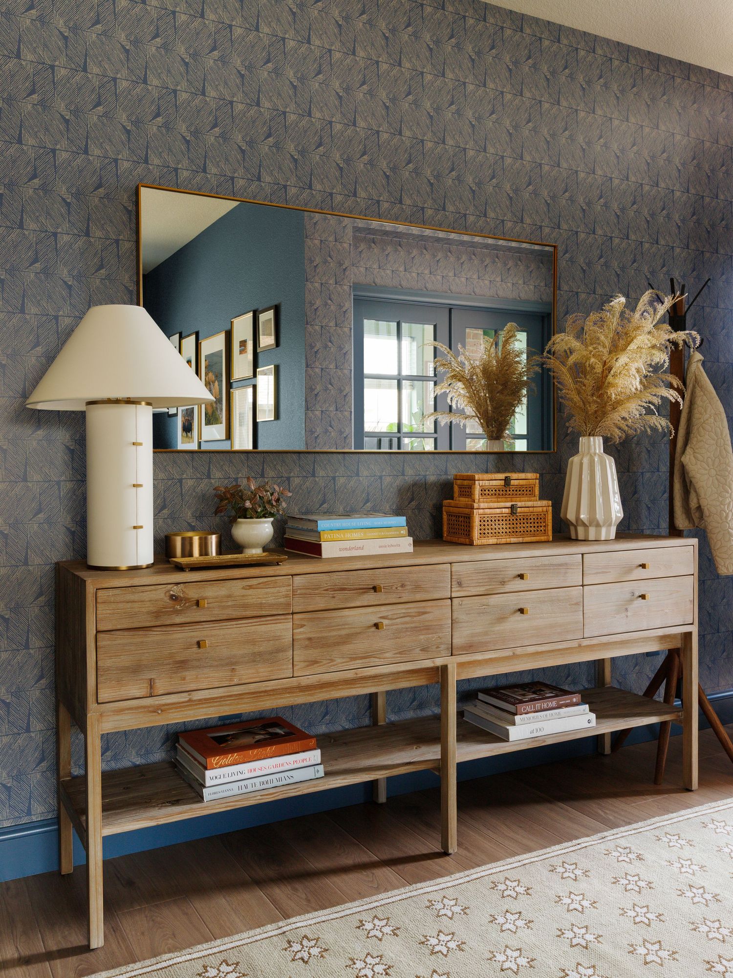 A wooden console dresser with drawers, styled with a lamp, vases, and books against blue floral wallpaper with a gold mirror.